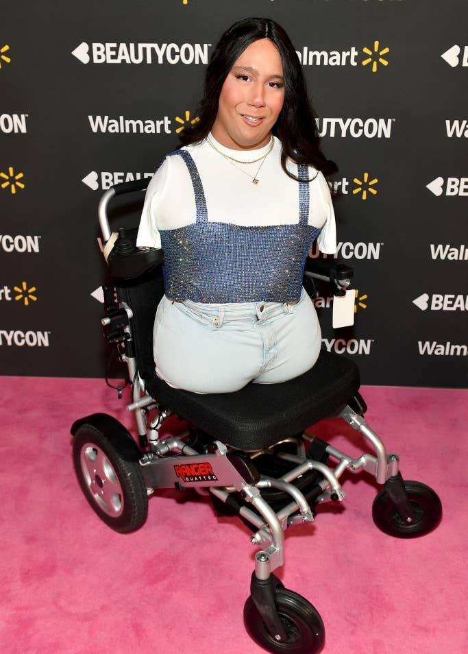 Person in a wheelchair on a pink carpet at an event, wearing a white top with a sequined overtop and light jeans. Sponsors' logos are in the background