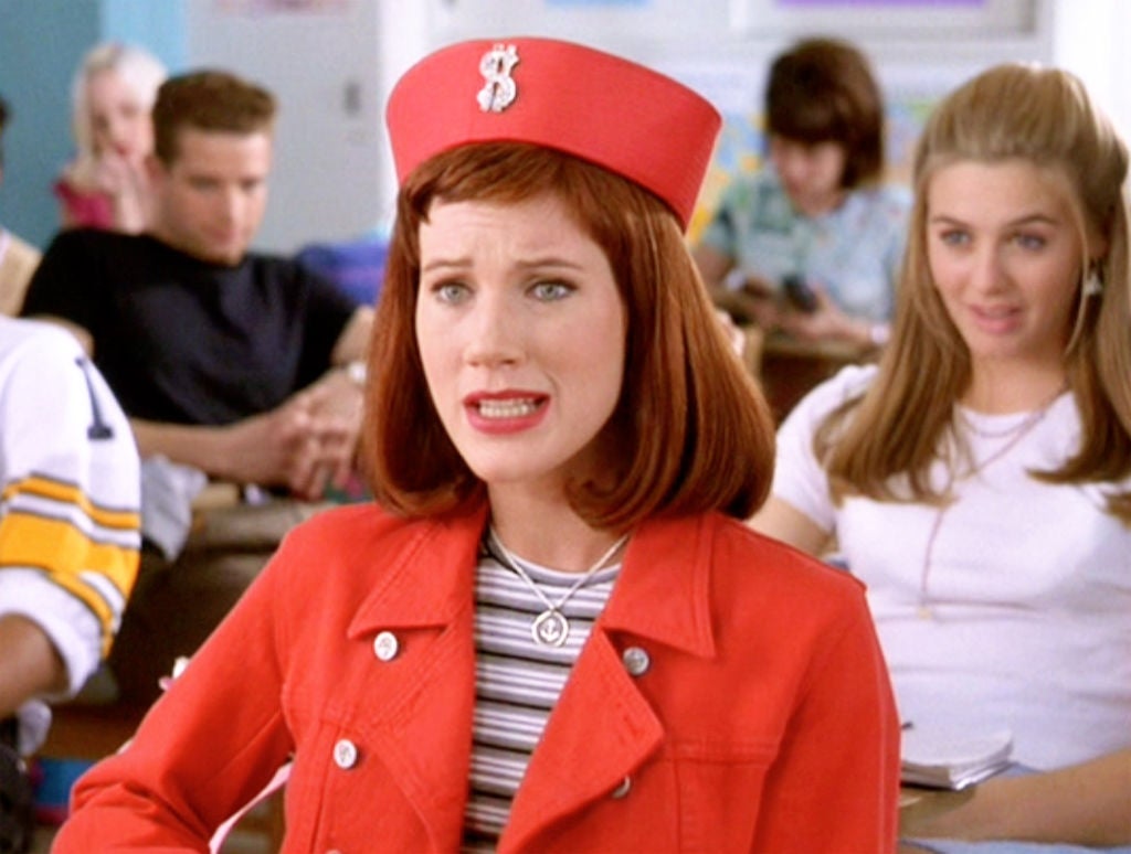 Person in a classroom wearing a retro-style outfit with a bright hat featuring a dollar sign, speaking animatedly. Others sit in the background