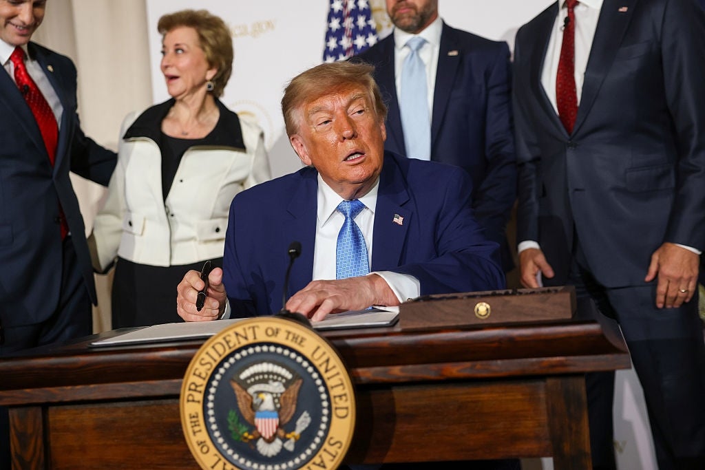 En person i dress sitter ved et skrivebord og signerer papirer, omgitt av andre i formell antrekk. Seglet til USAs president er synlig