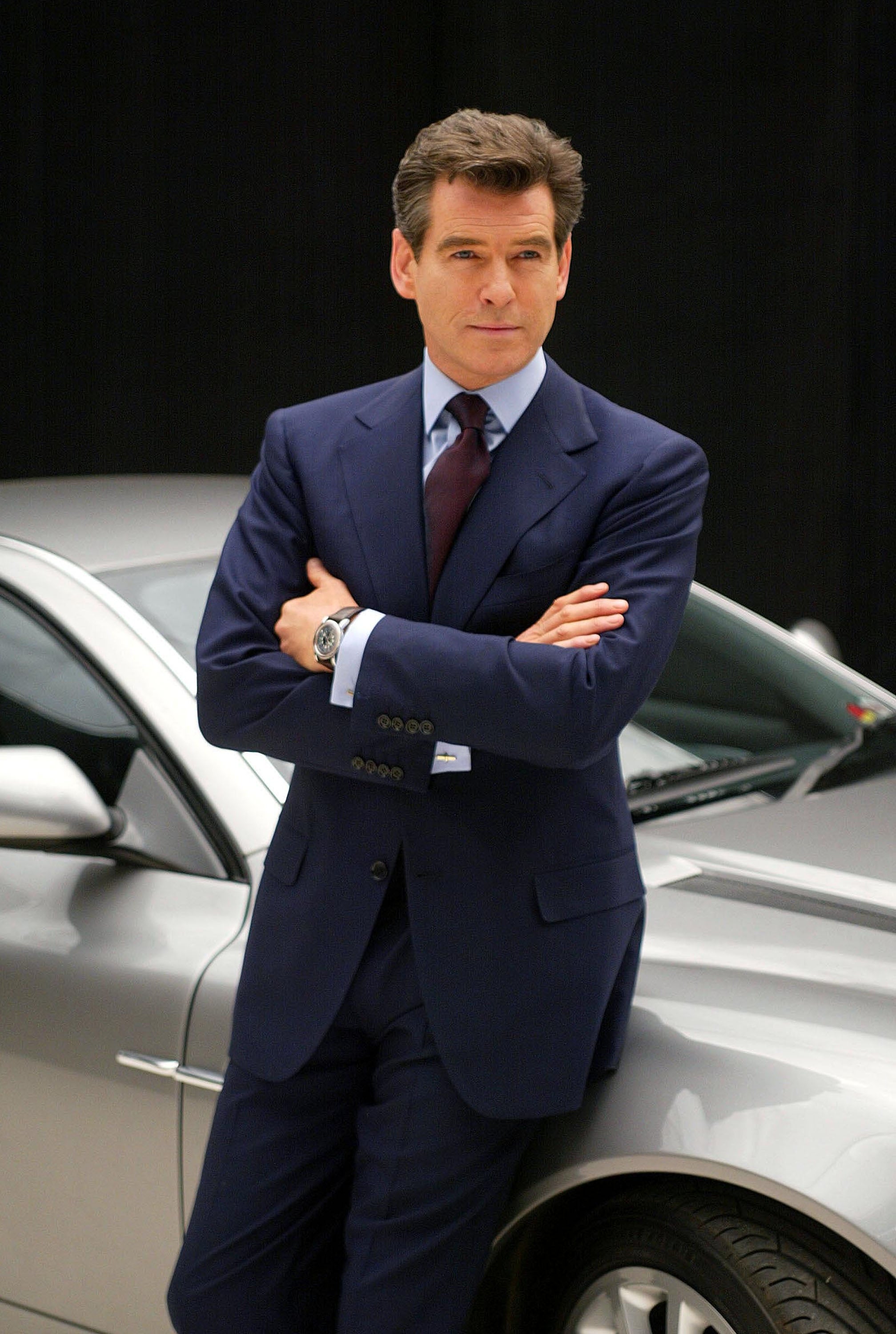 Man in a suit leans confidently against a luxury car with arms crossed, conveying a sophisticated and composed demeanor
