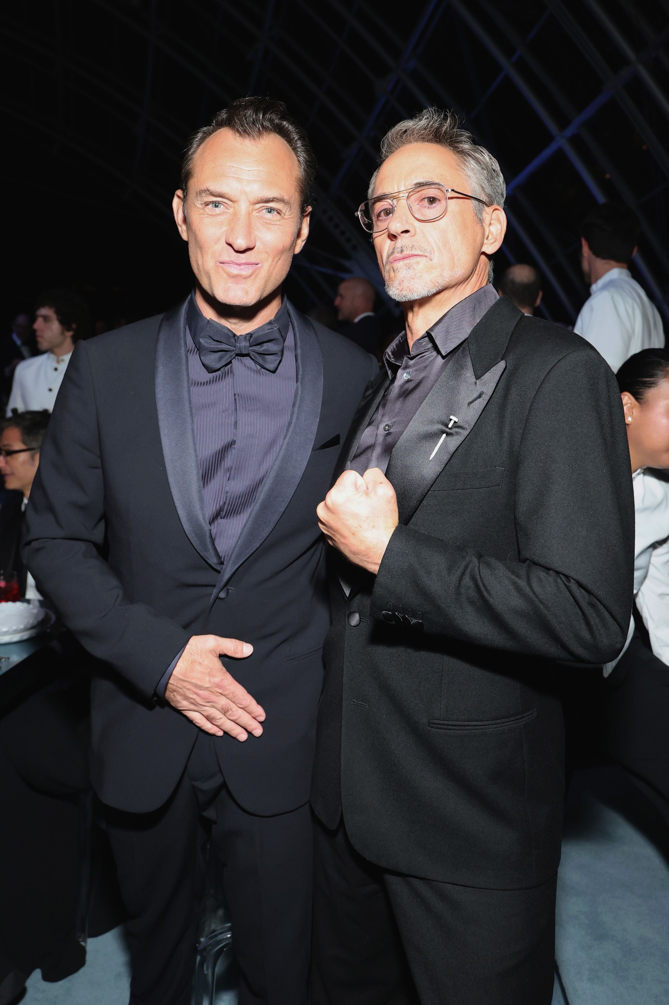 Two men in stylish black suits pose confidently at a formal event, surrounded by attendees in the background