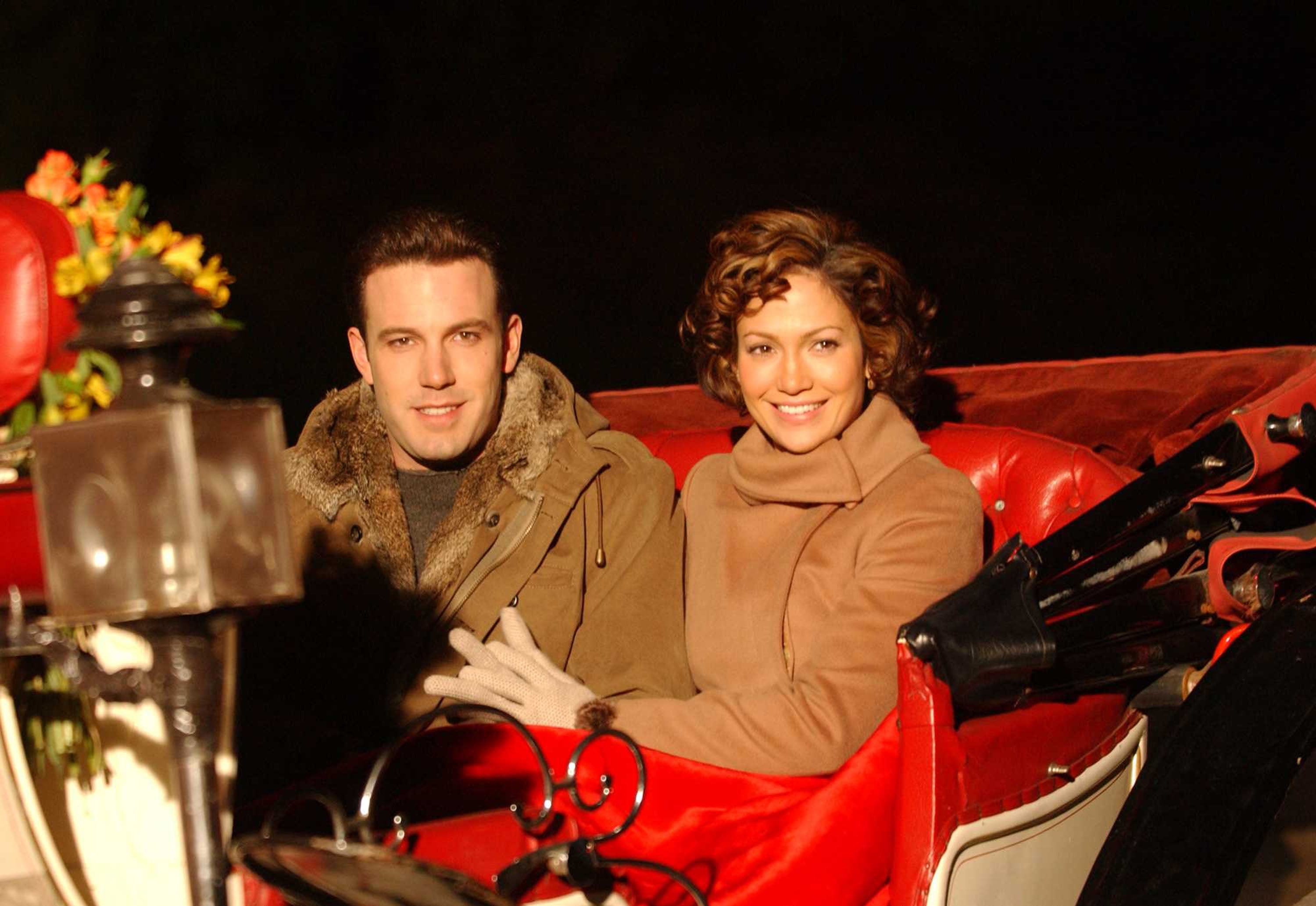 Two people in winter coats smile while sitting in a horse-drawn carriage, appearing happy and cozy at a nighttime event