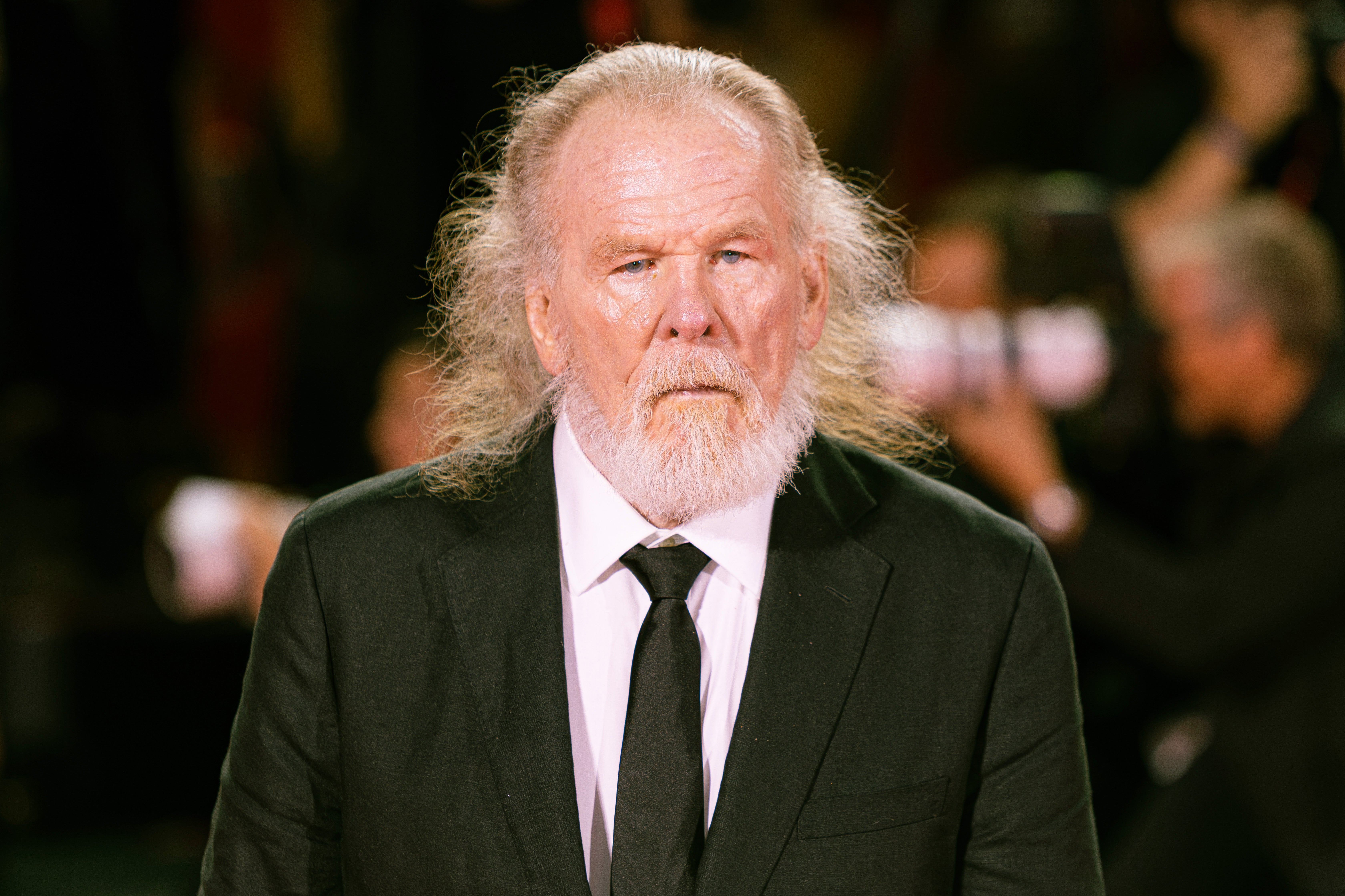 An older man with long white hair and a beard wears a classic black suit and tie on a red carpet, surrounded by photographers