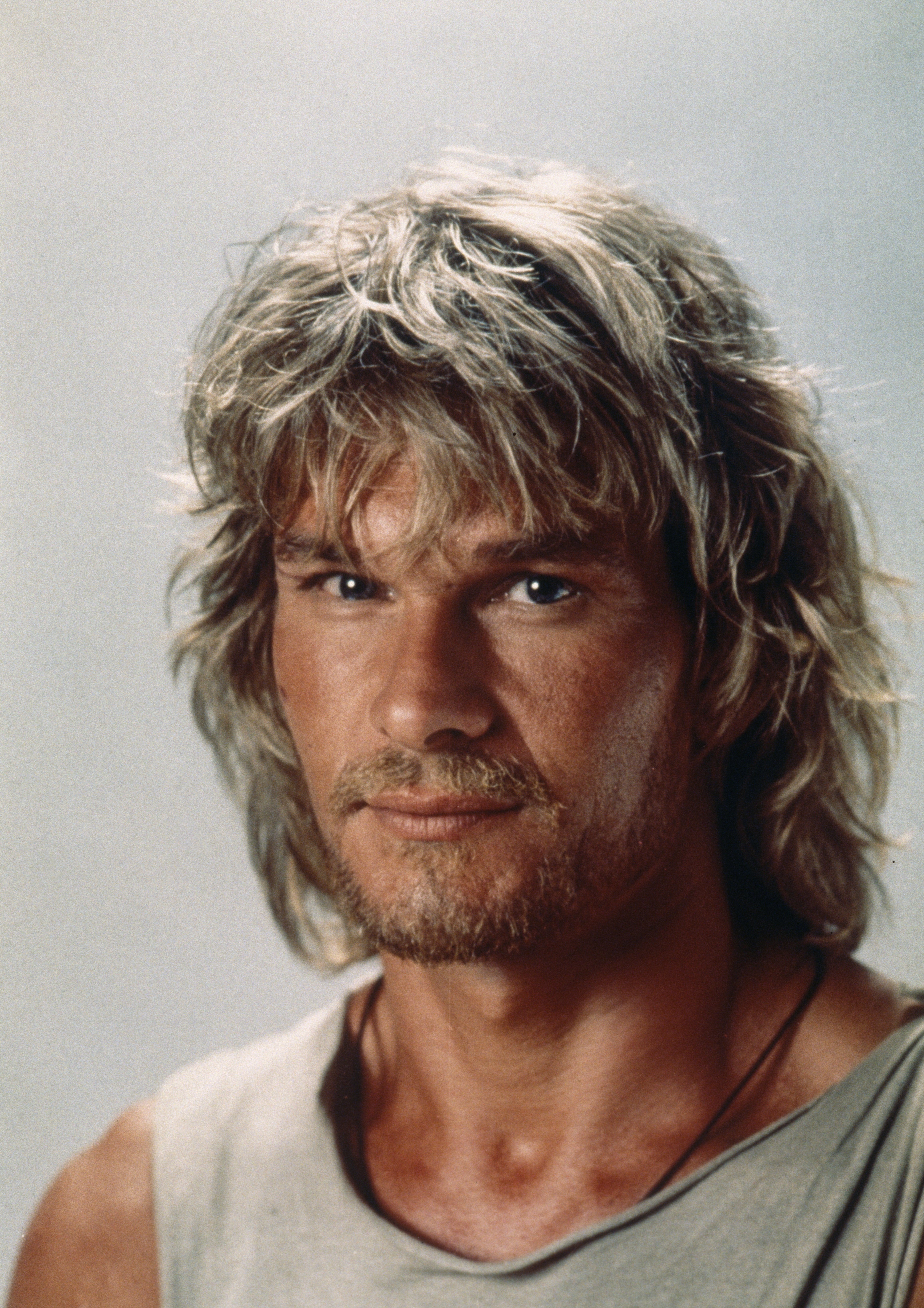 Person with shaggy hair and a beard, wearing a sleeveless top, looking directly at the camera