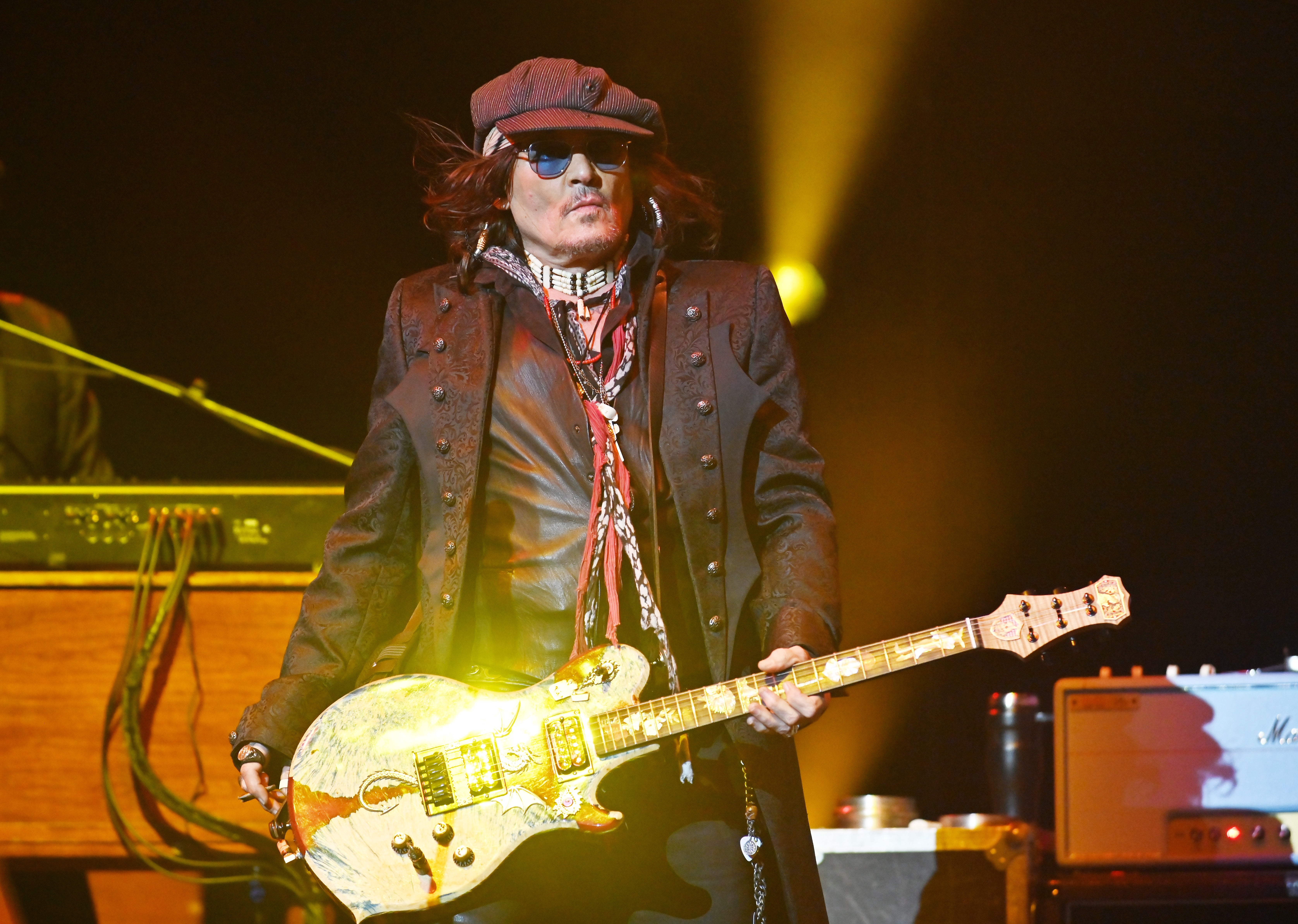 Musician playing electric guitar on stage, wearing a layered outfit with a jacket, hat, and sunglasses