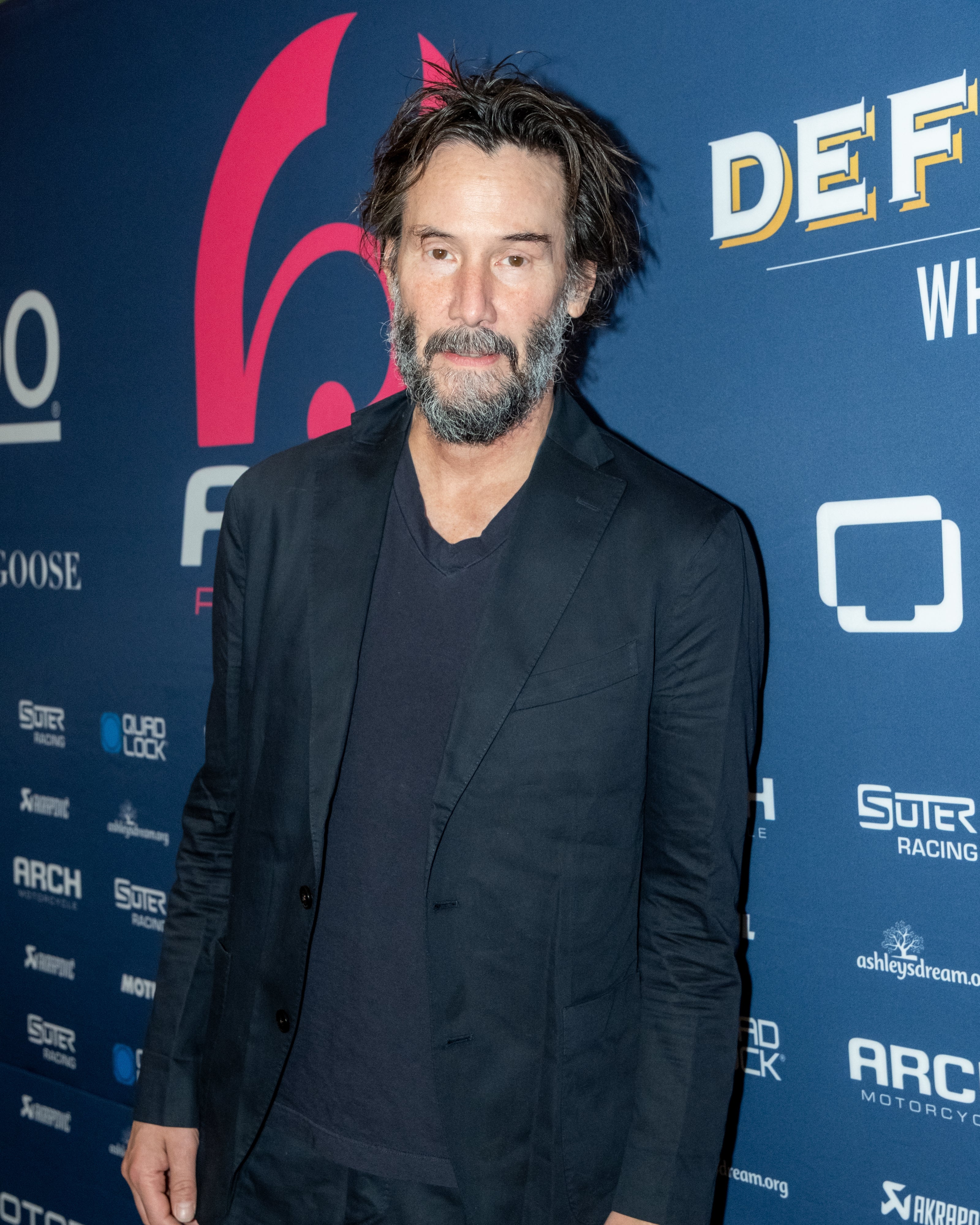 Person in a casual suit on a red carpet poses calmly at an event backdrop