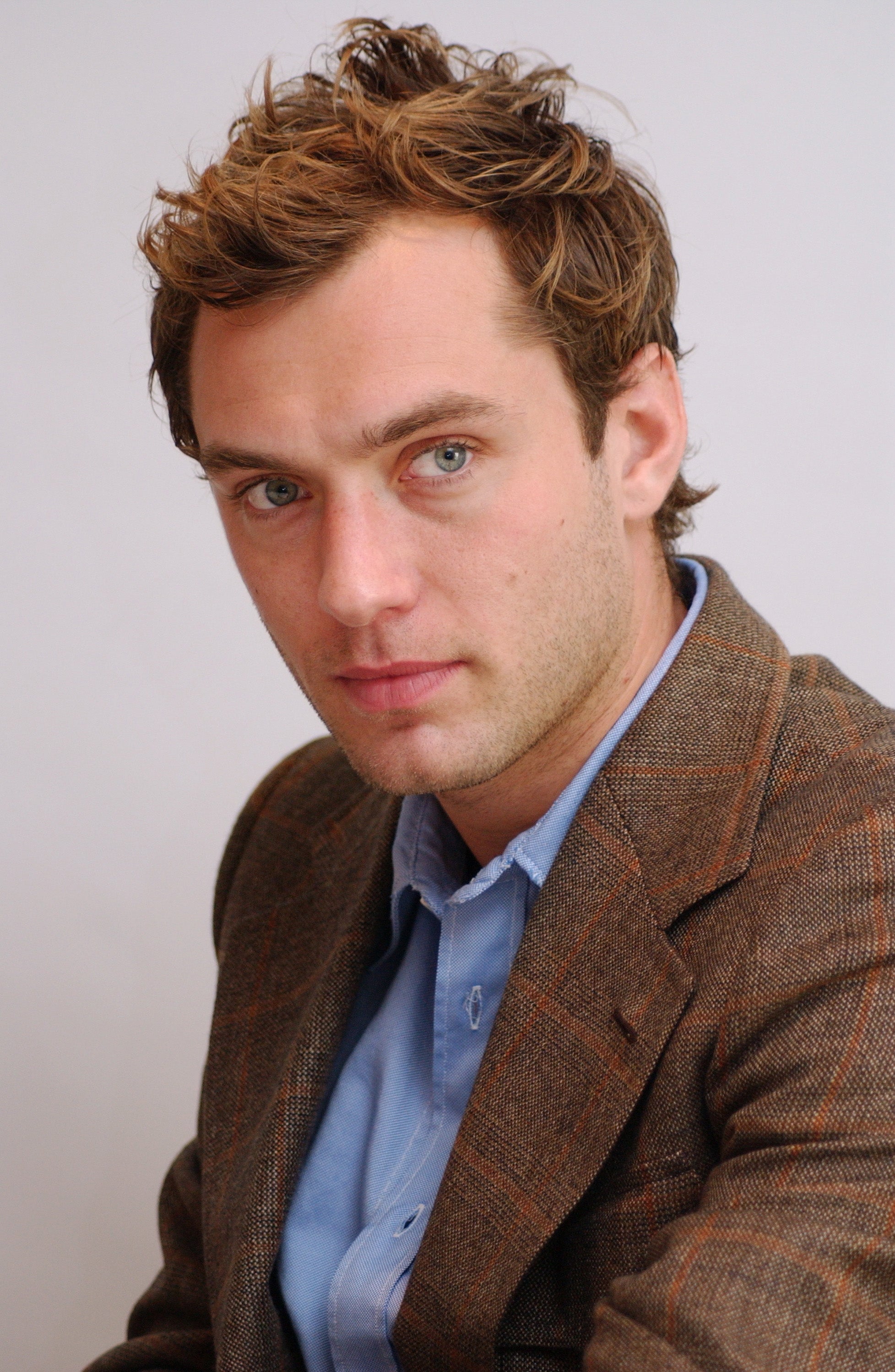 A person with styled hair wears a tailored plaid suit and a blue shirt, gazing directly at the camera