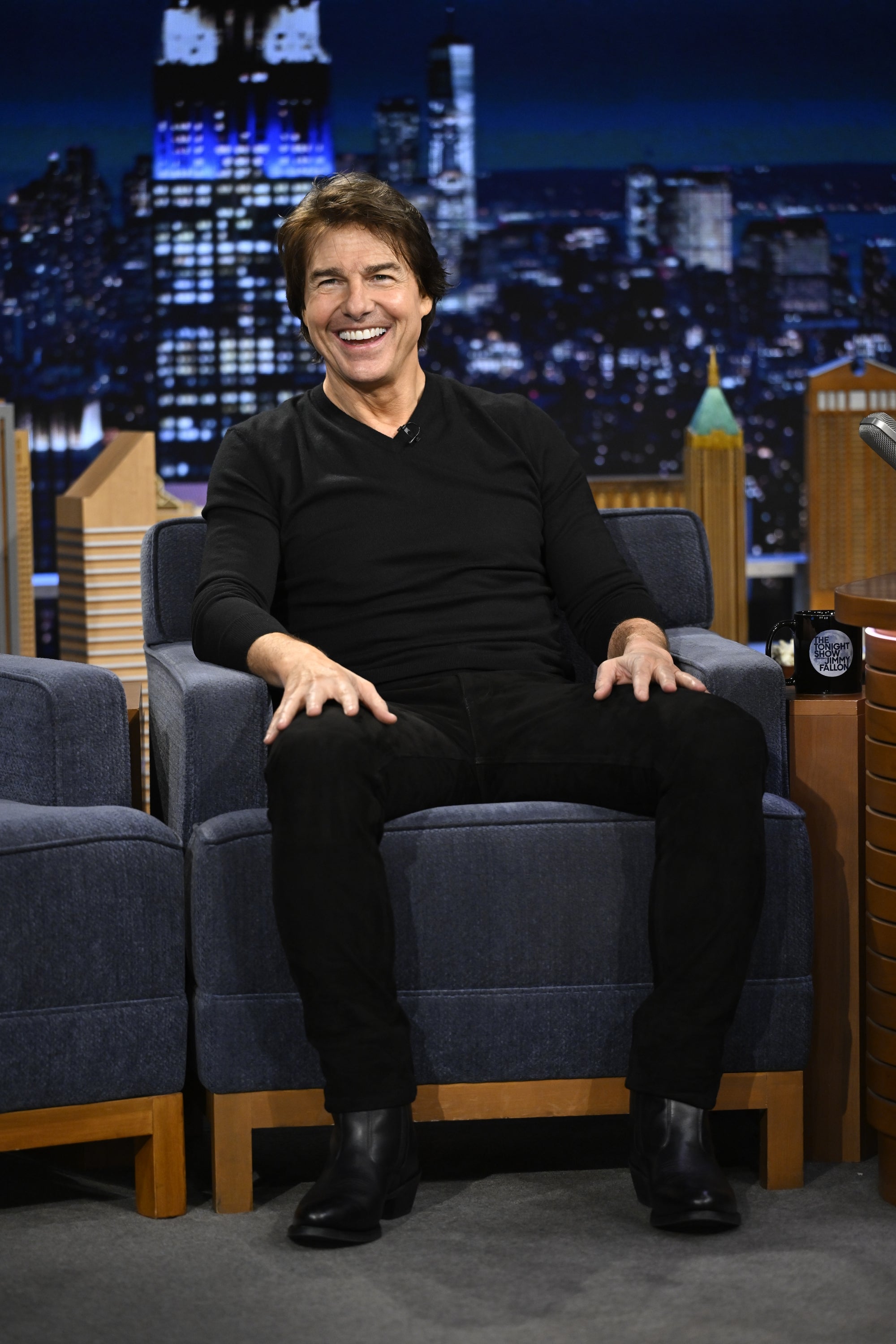 Actor sits on a talk show set, smiling, in casual black attire and boots. Background shows a cityscape display