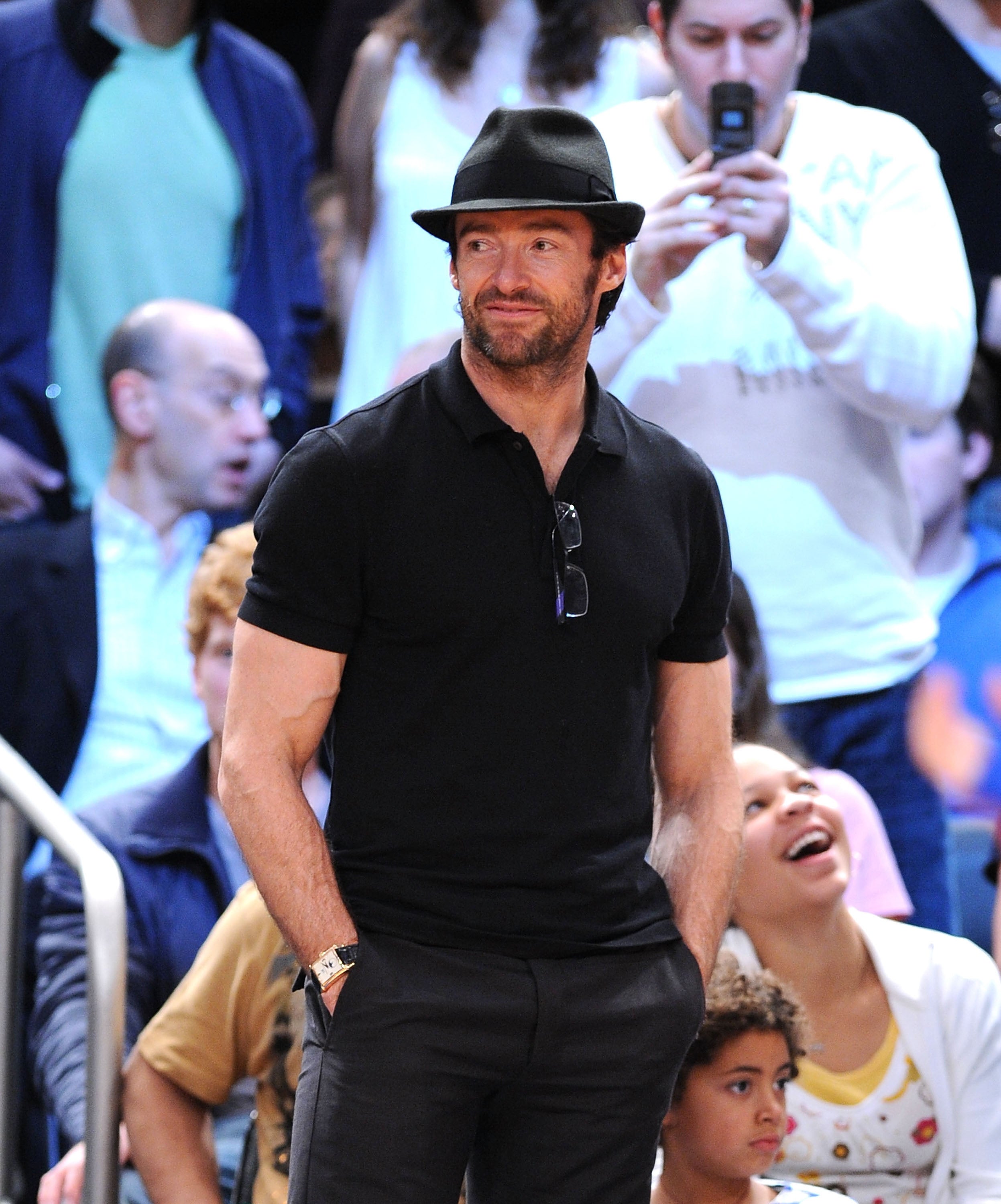 Man in a black polo and fedora stands casually with hands in pockets, smiling among a crowd at an event