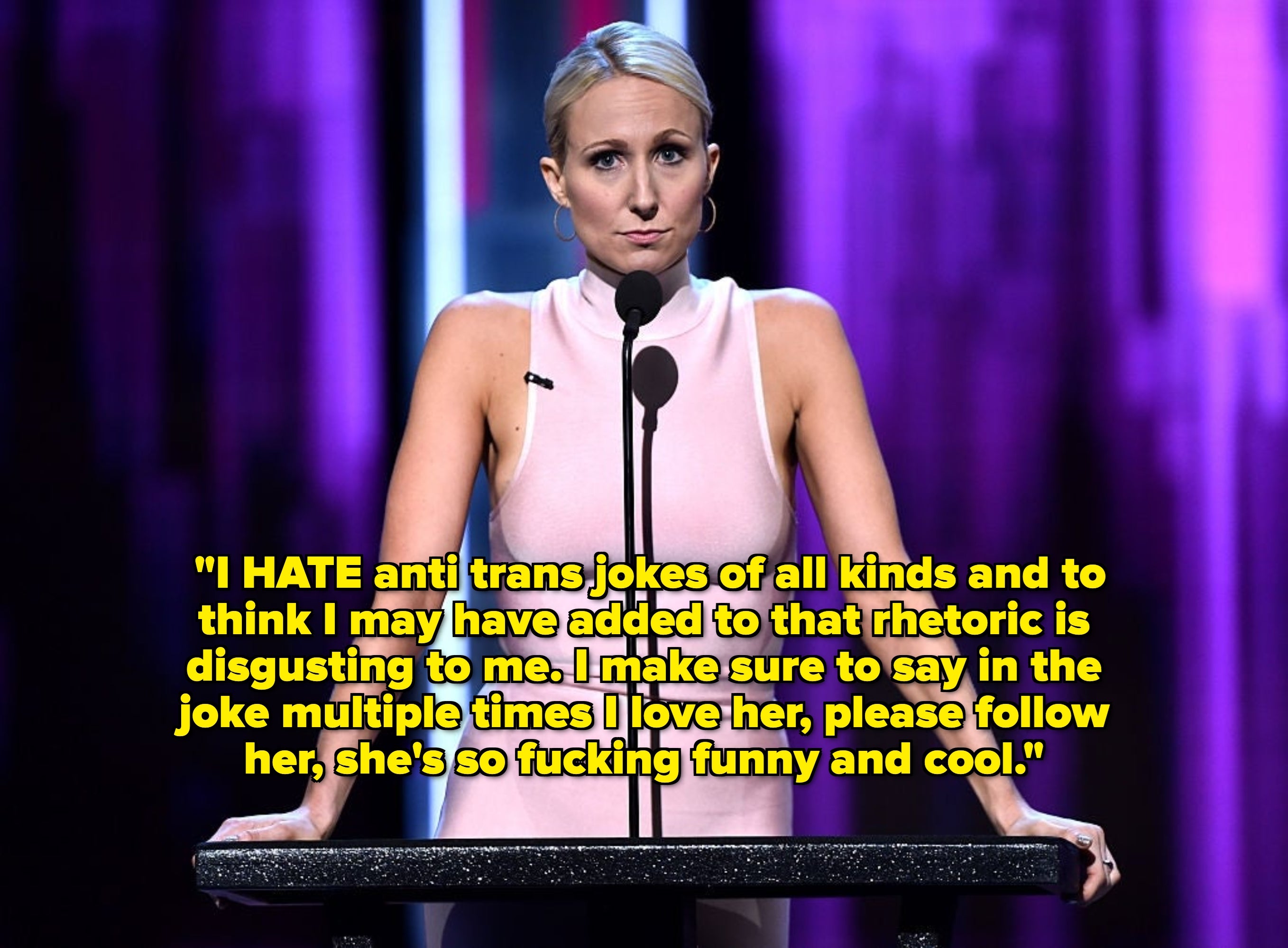 Person in a sleeveless dress stands at a podium on stage, speaking into a microphone during a public event