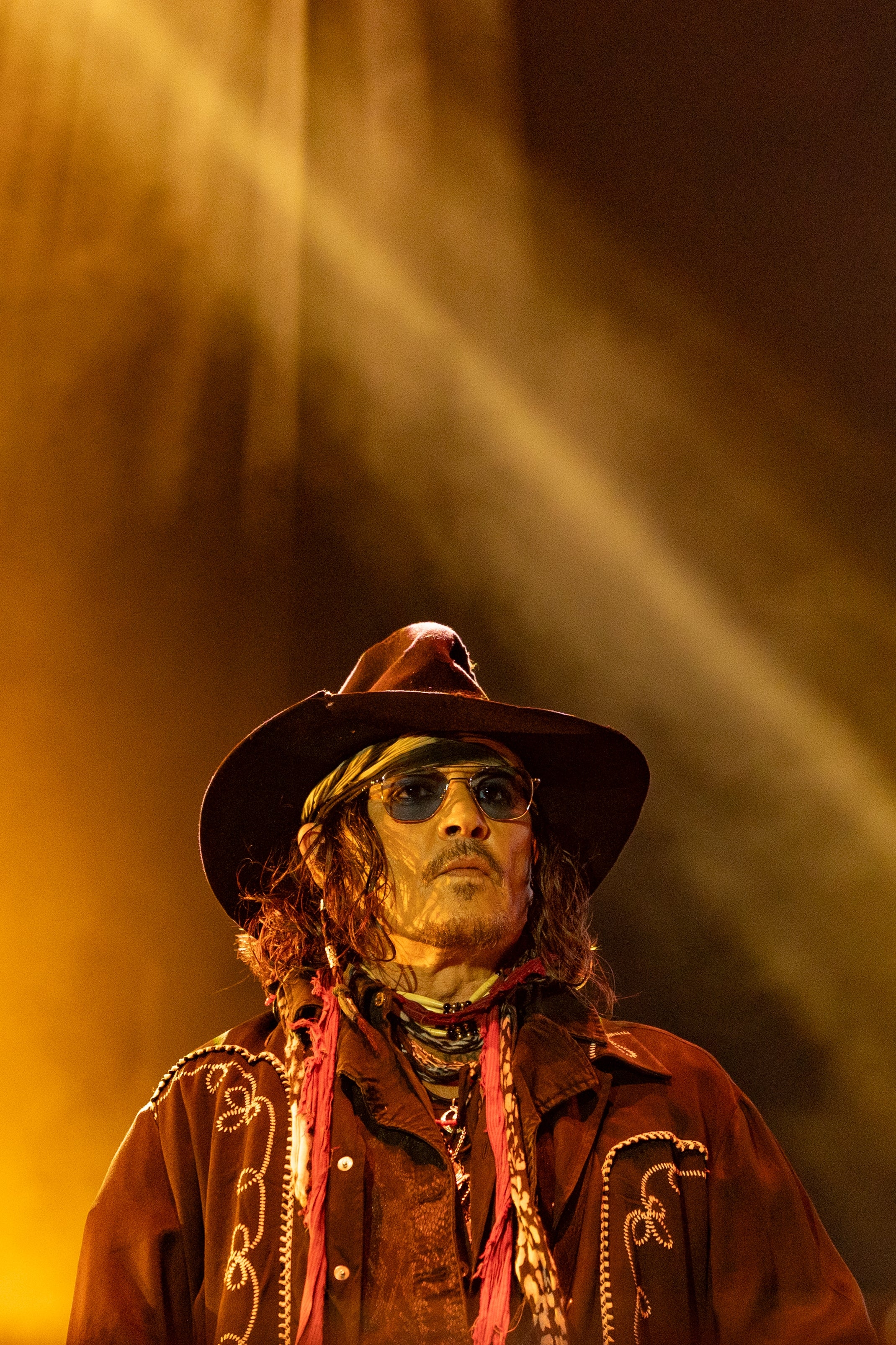 Person wearing a wide-brimmed hat, sunglasses, and an embroidered jacket, standing under stage lights