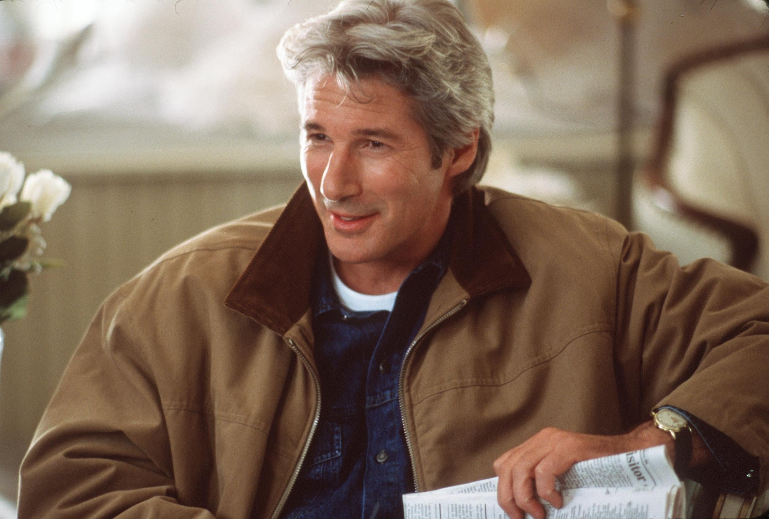 A person in a casual jacket and shirt, seated, holding a newspaper