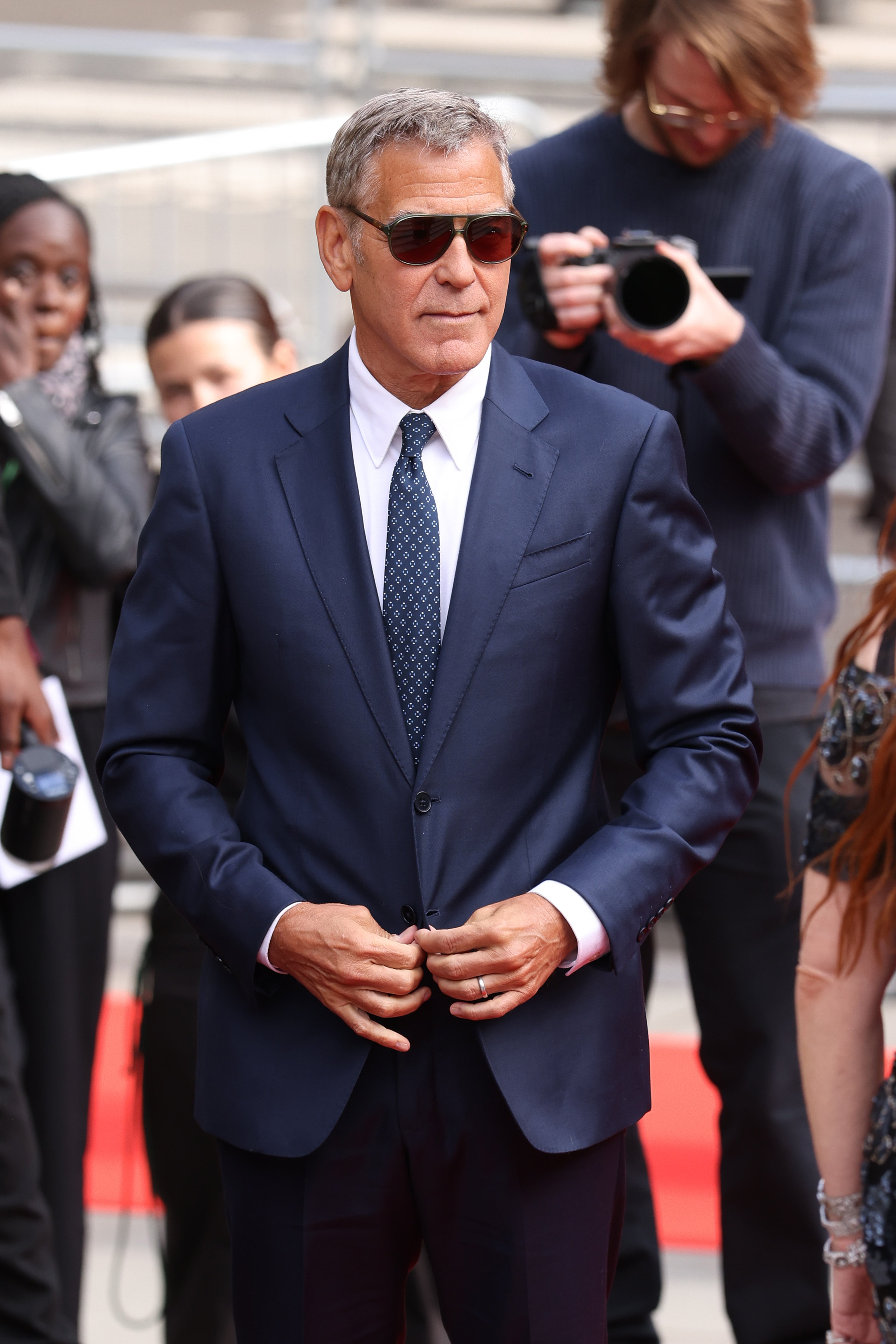 Man in a tailored suit and sunglasses on a red carpet, surrounded by event attendees and photographers