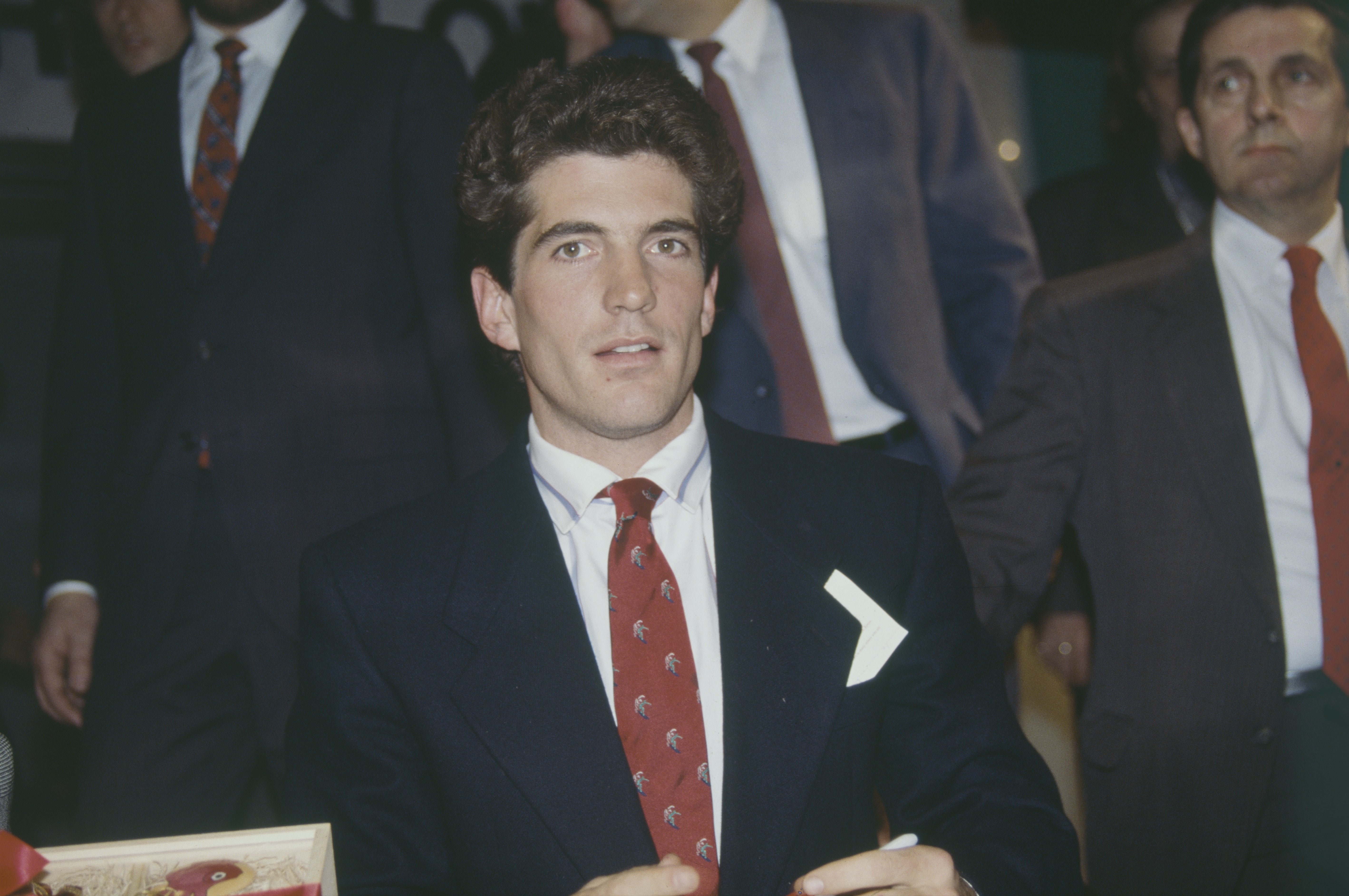 A man in a suit with a patterned tie stands at an event, surrounded by others in formal attire