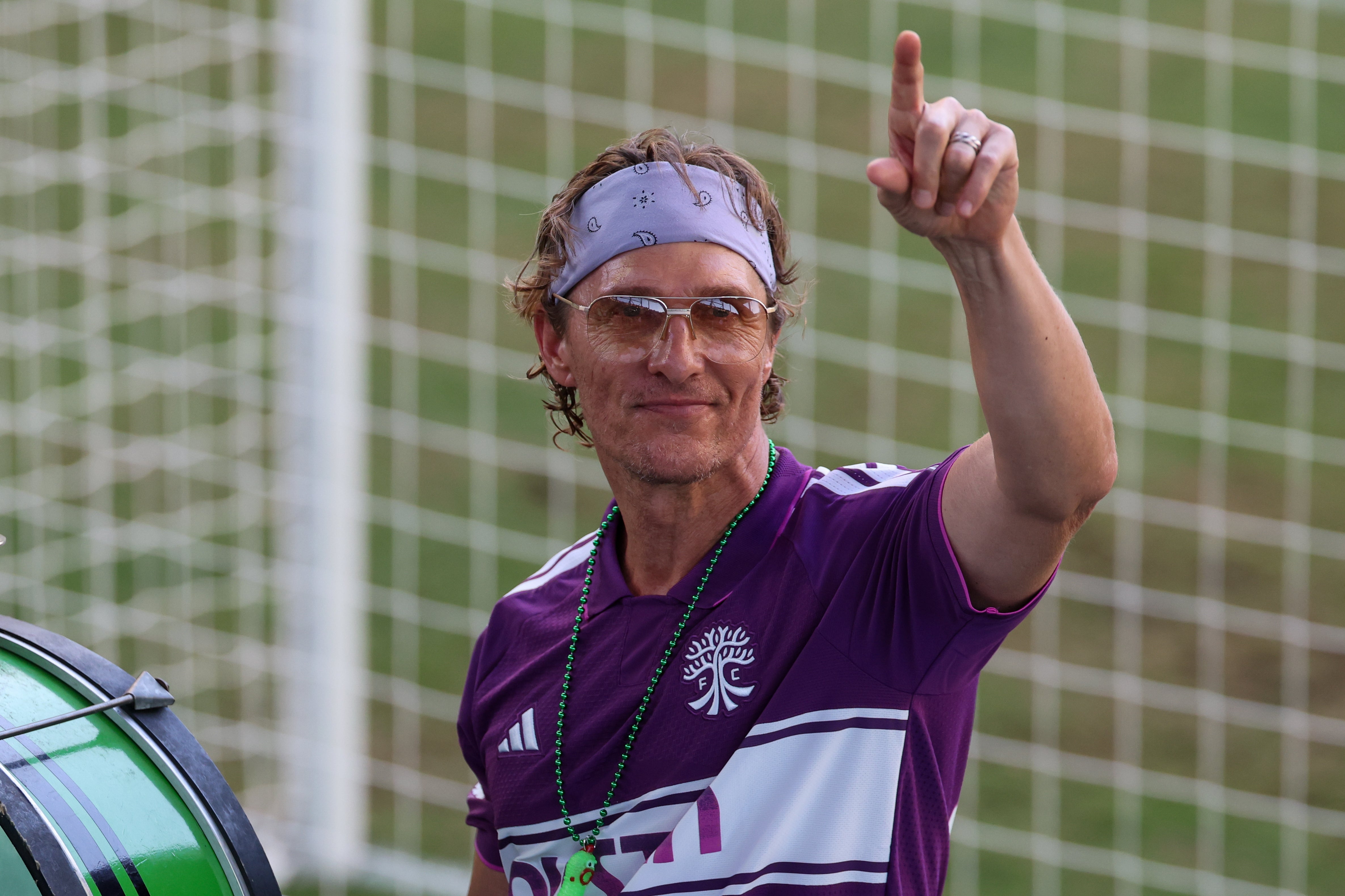 A person in a casual sporty outfit with a headband gestures with one hand, standing in front of a soccer goal