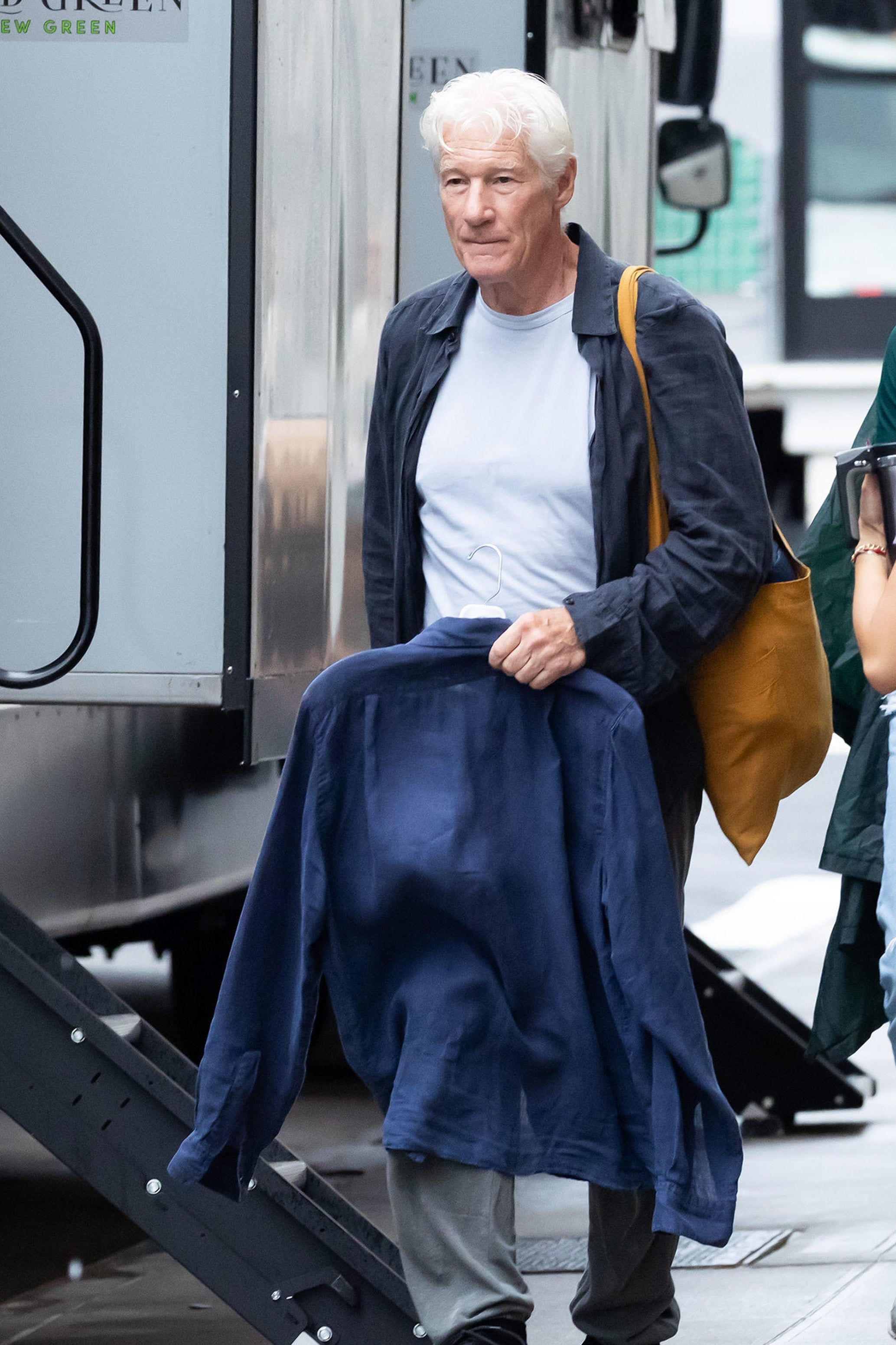 An unidentified man in casual attire, holding a jacket and a tan bag, walks near a trailer, suggesting a movie set or event preparation