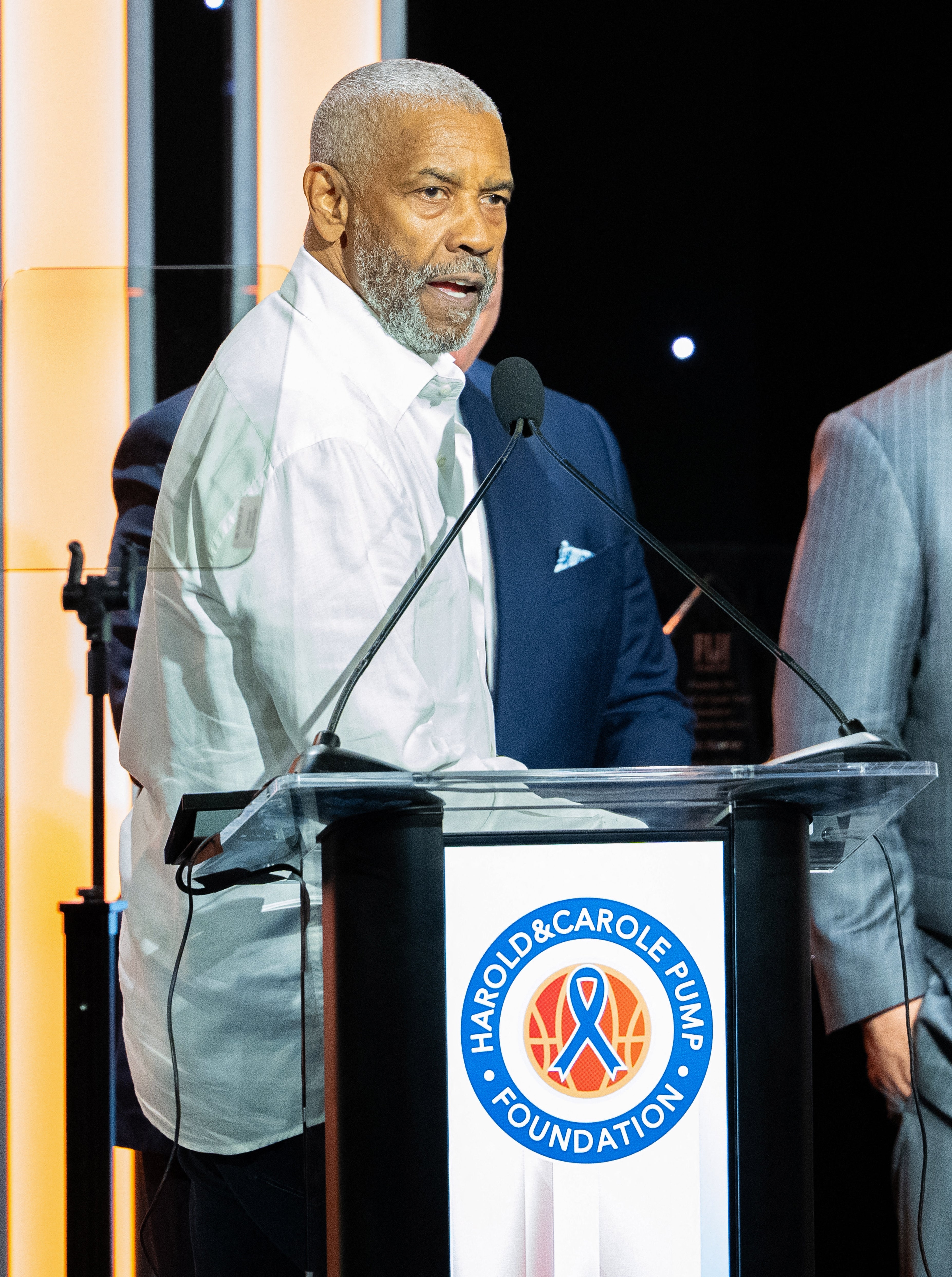 Man in a white shirt speaks at a podium during an event. The podium has a sign for the Harold &amp;amp; Carole Pump Foundation
