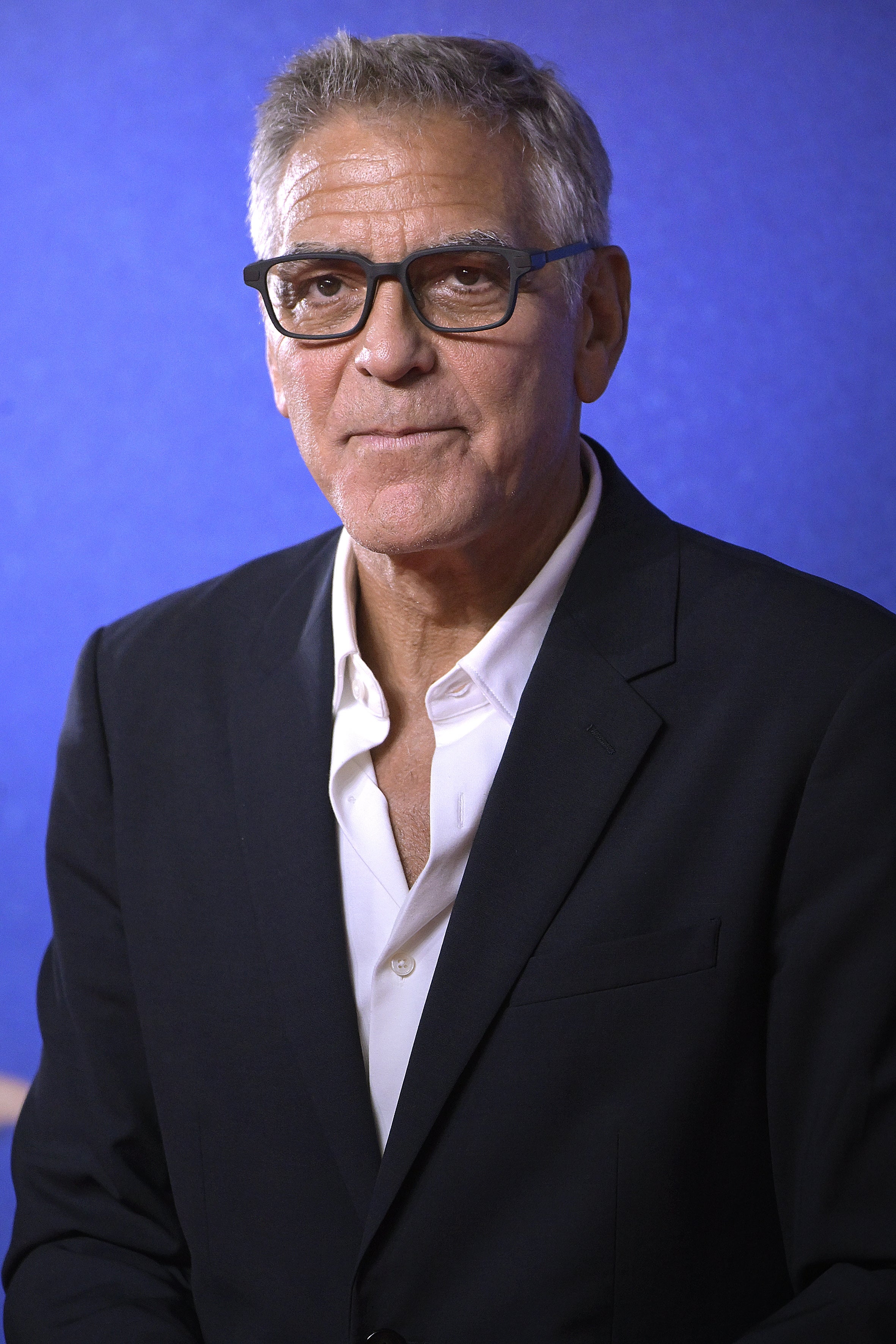 A man in a blazer and open-collared shirt poses in front of a blue background