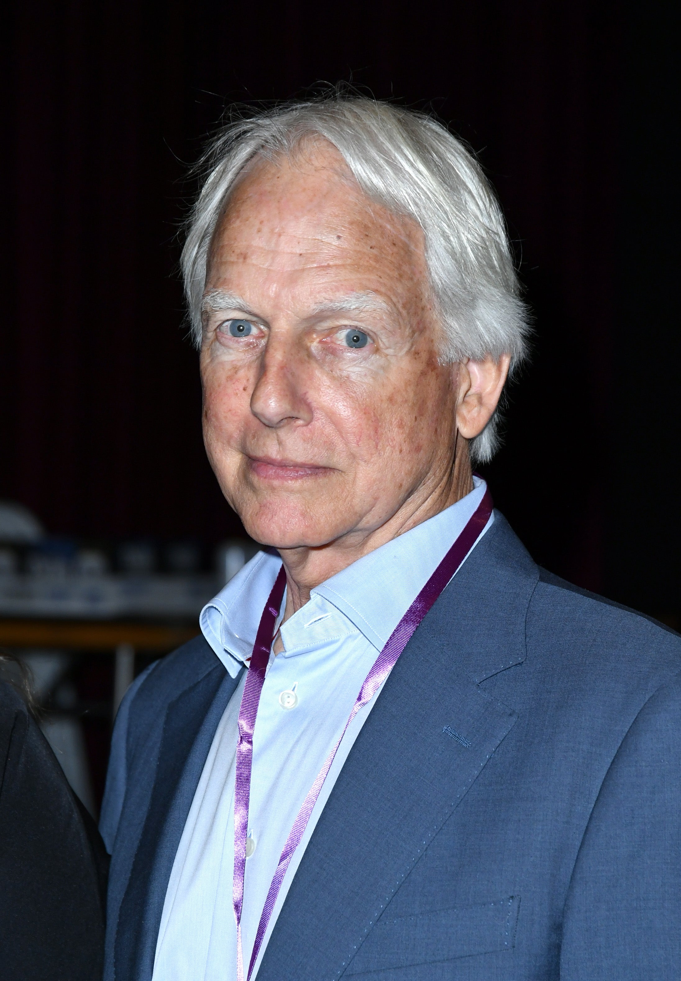 Elderly man in a suit with a purple lanyard, posing on a red carpet event
