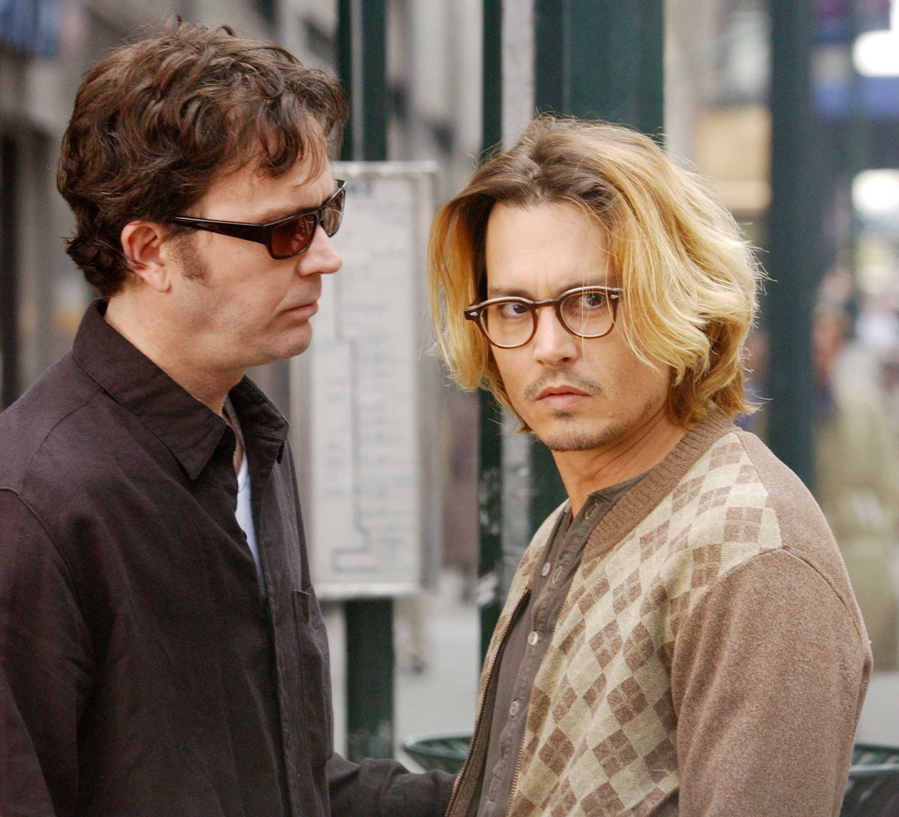 Two individuals in casual clothing with glasses, in a city street setting. One has short hair, the other has medium-length hair
