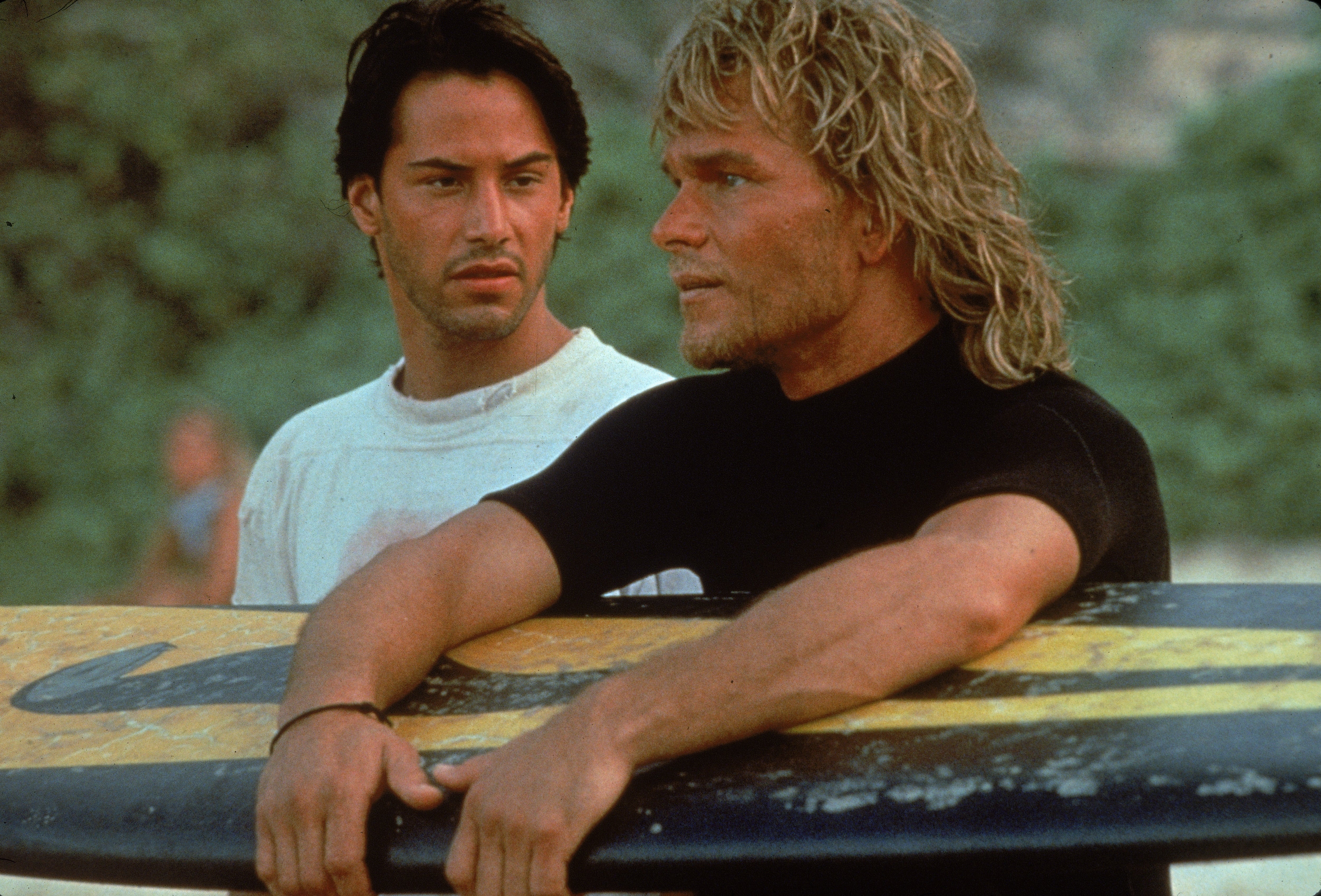Two men stand by a surfboard on a beach. One wears a wetsuit, the other a casual shirt. They appear focused on something in the distance