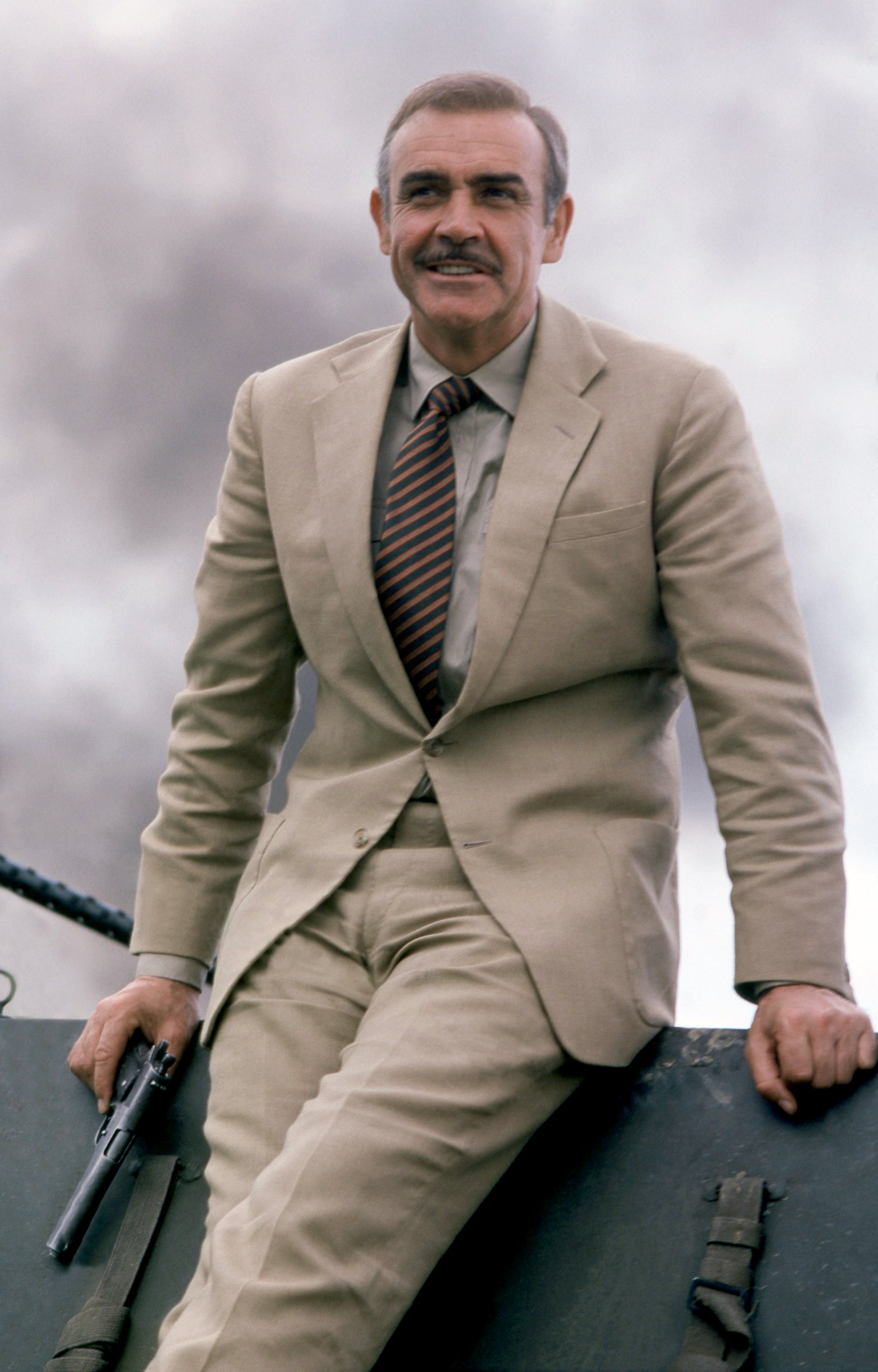 Person in a classic suit with a striped tie, sitting outdoors, holding a gun, against a backdrop of smoke