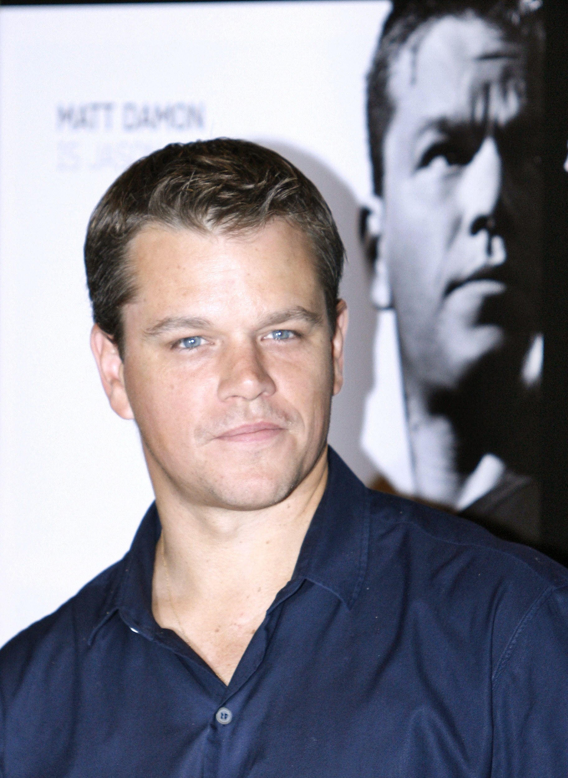 Person in button-up shirt at an event, posing in front of their black-and-white portrait