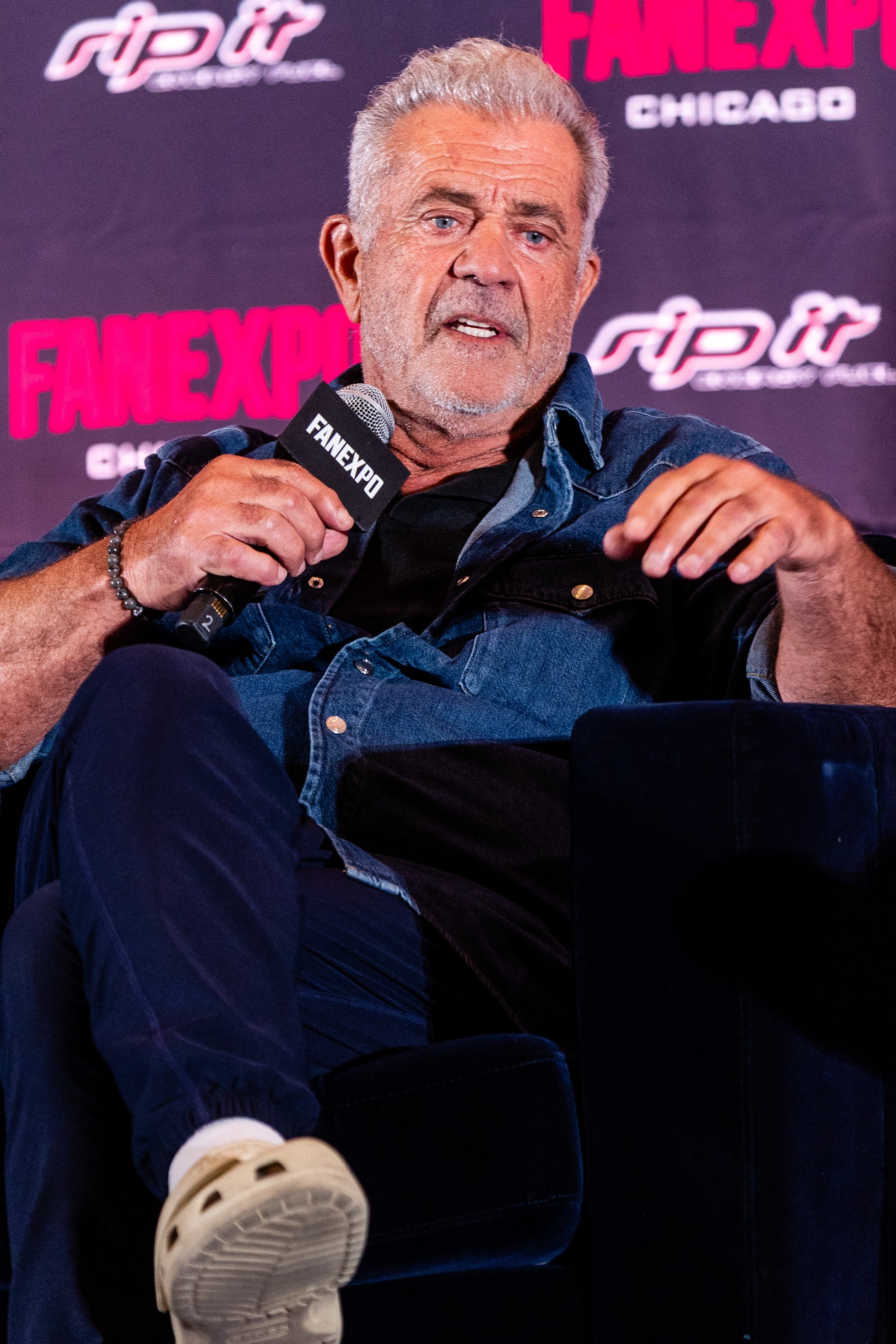 A person sits on stage at a fan expo event, holding a microphone and wearing casual jeans and a denim shirt