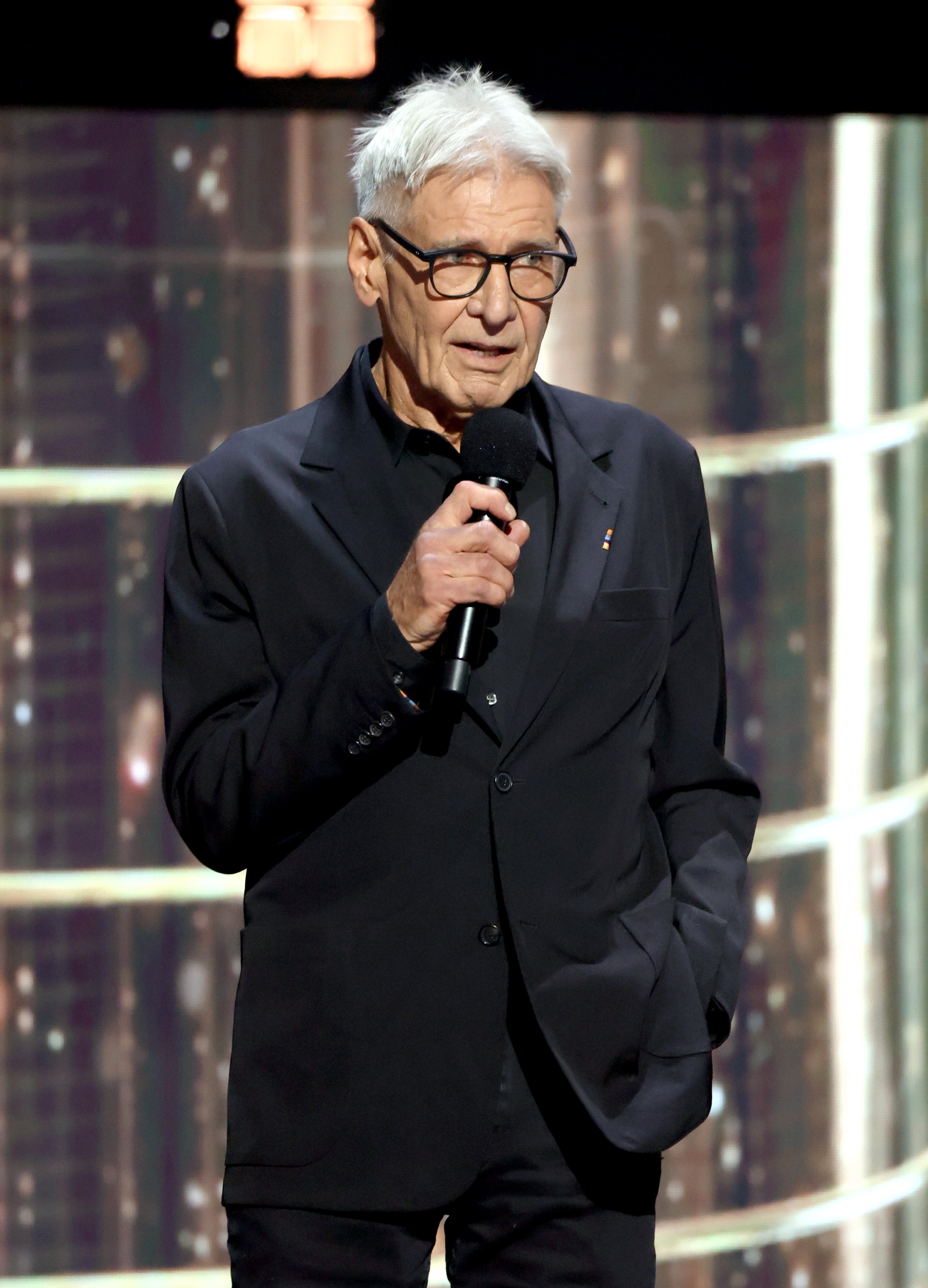Person in a black suit speaks into a microphone on stage at a formal event