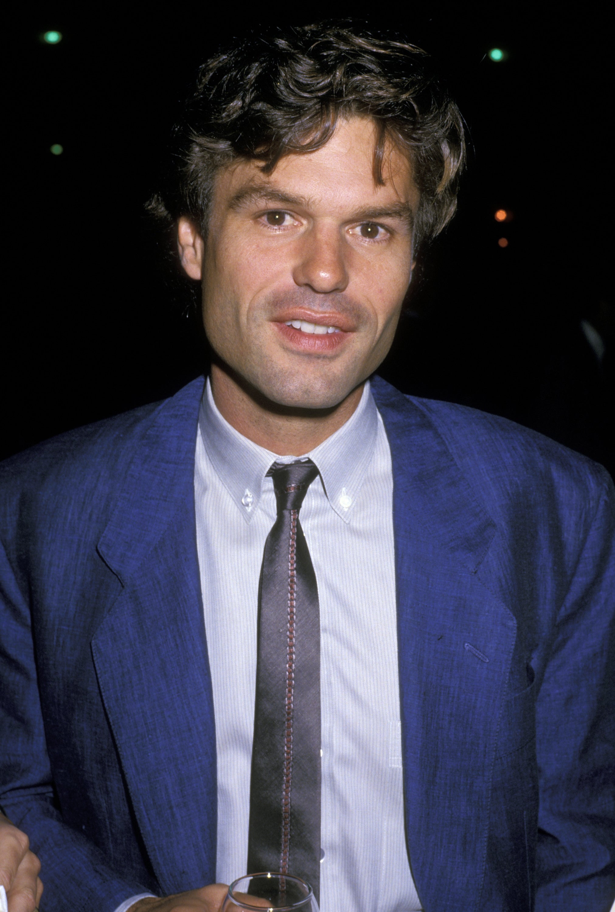 A person in a suit with a relaxed hairstyle, wearing a tie, attends an event, holding a drink