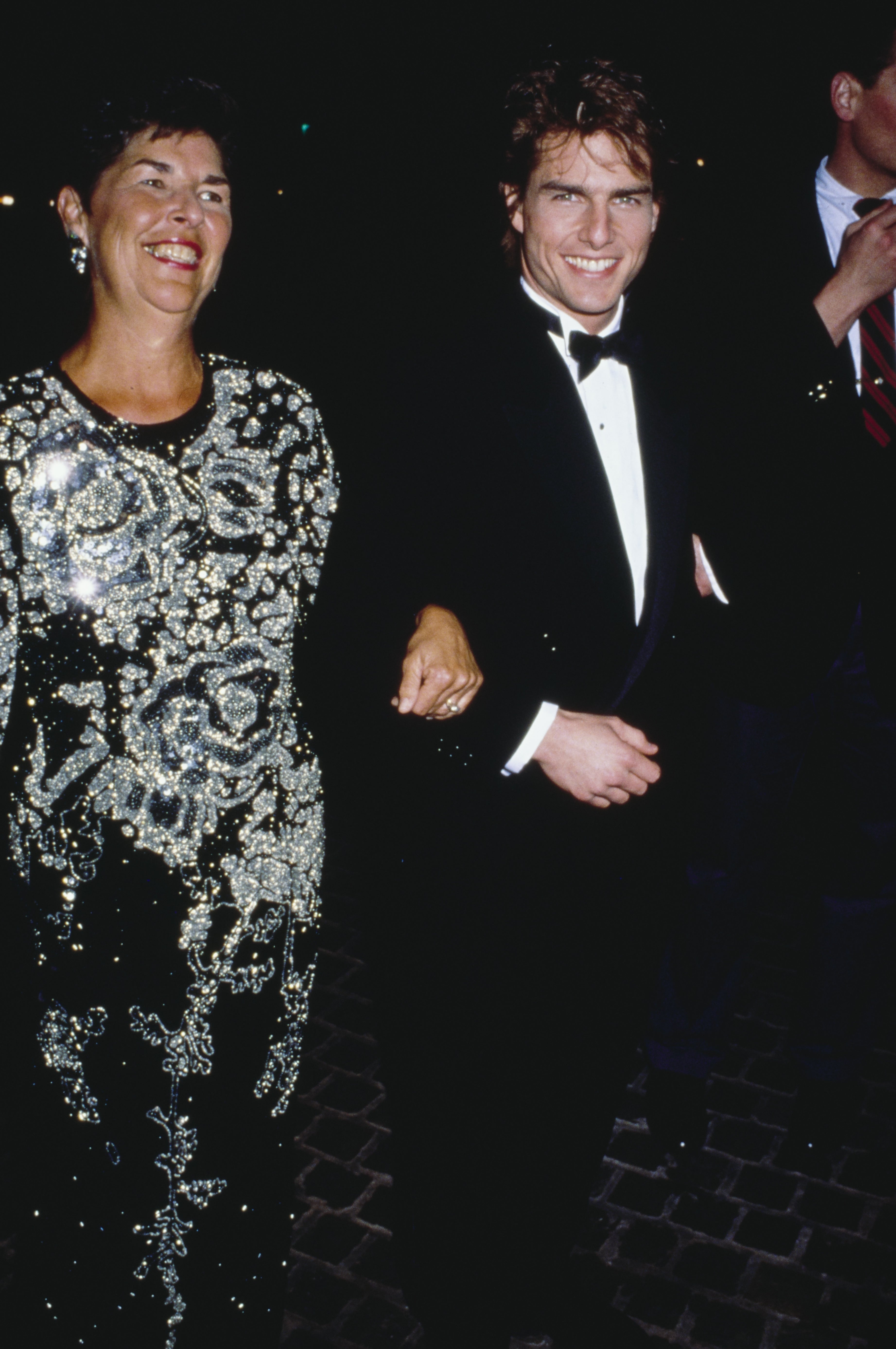 A man in a tuxedo smiles while linking arms with a woman in a sequined dress at an event