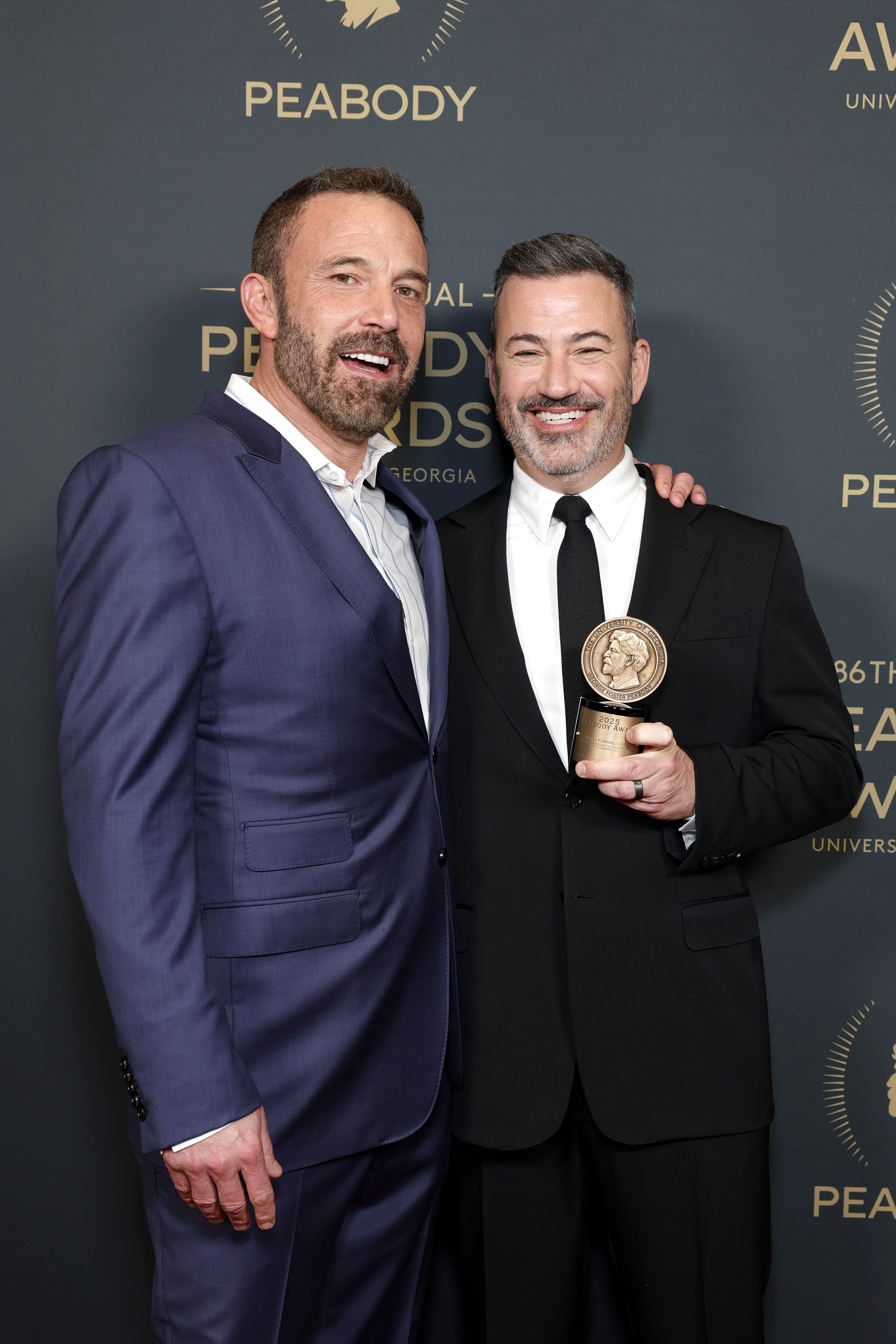 Two people in formal attire at an awards event, one holding a trophy and both smiling in front of a backdrop with "Peabody Awards" text