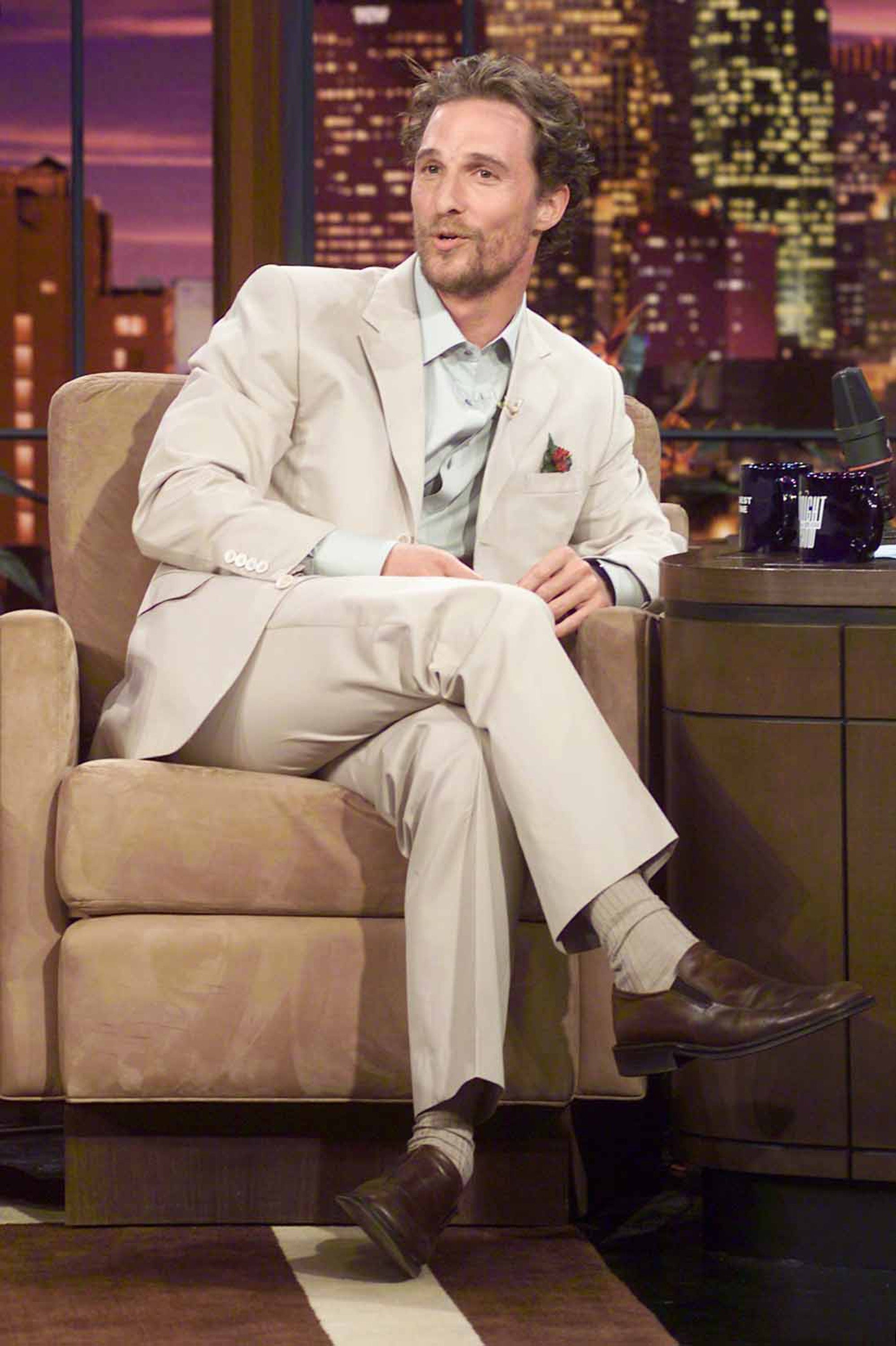 Actor in a light suit sits cross-legged on a talk show set, engaged in conversation