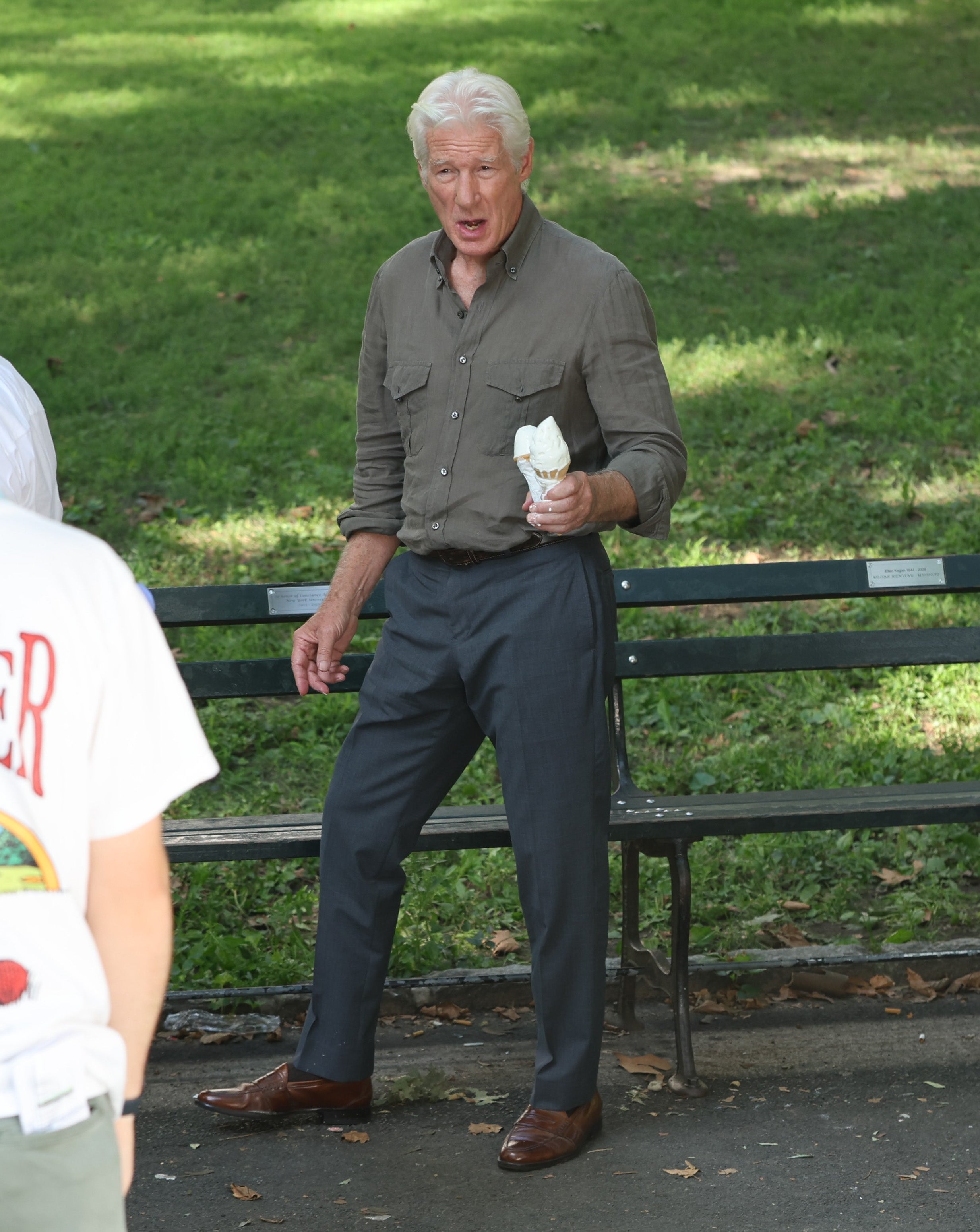 An older man stands in a park holding an ice cream cone, wearing a casual button-up shirt and trousers