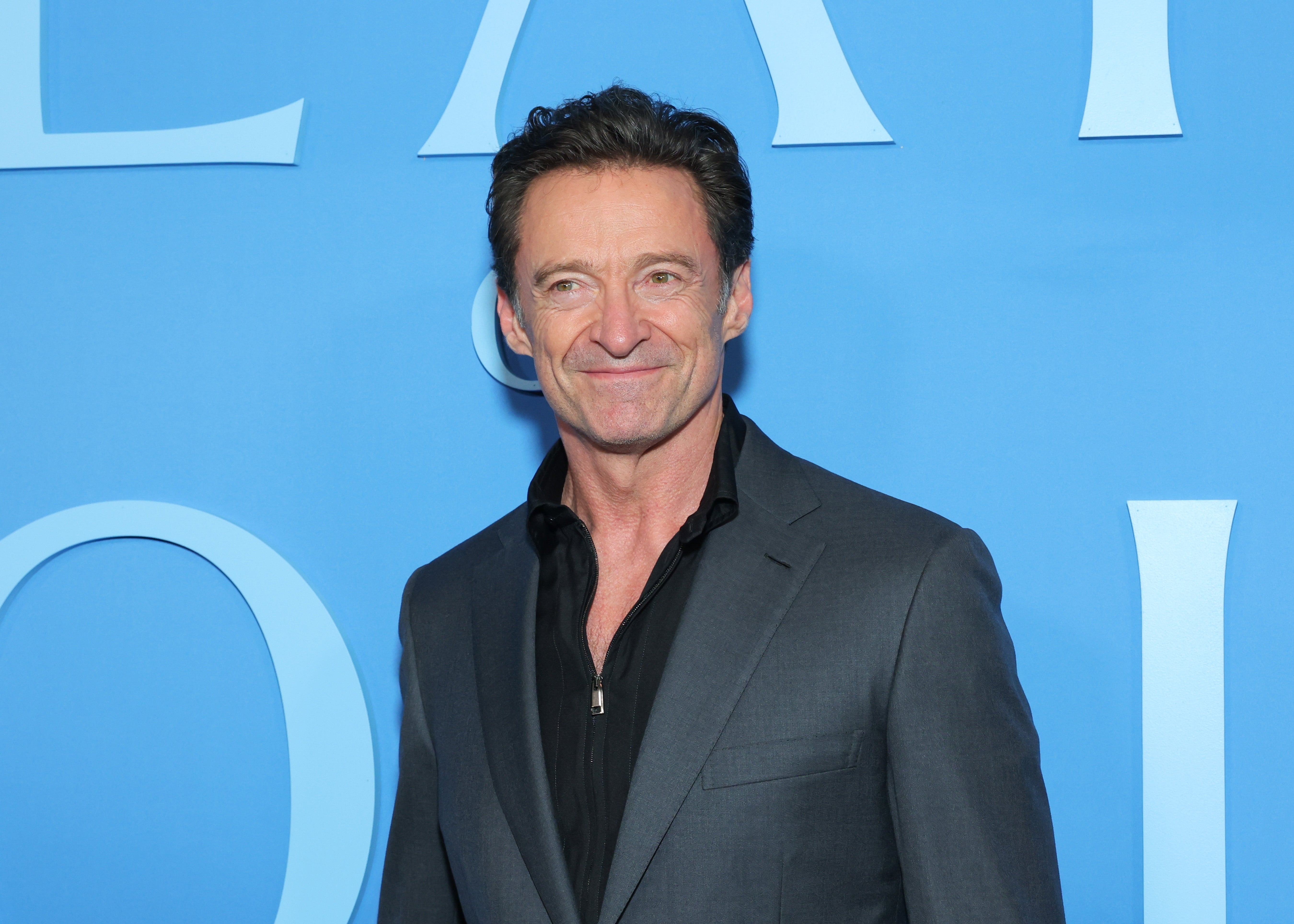 A person in a dark suit with a black shirt poses at a formal event against a blue backdrop with large letters