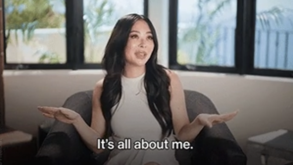 Woman in a white sleeveless dress sits on a chair, gesturing with her hands. Caption reads: "It's all about me."