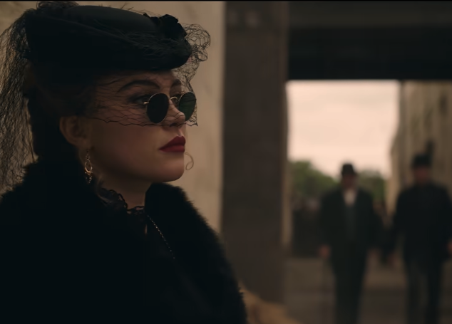 Person wearing a classic, vintage-style outfit with a black veil hat and round sunglasses, standing in a corridor