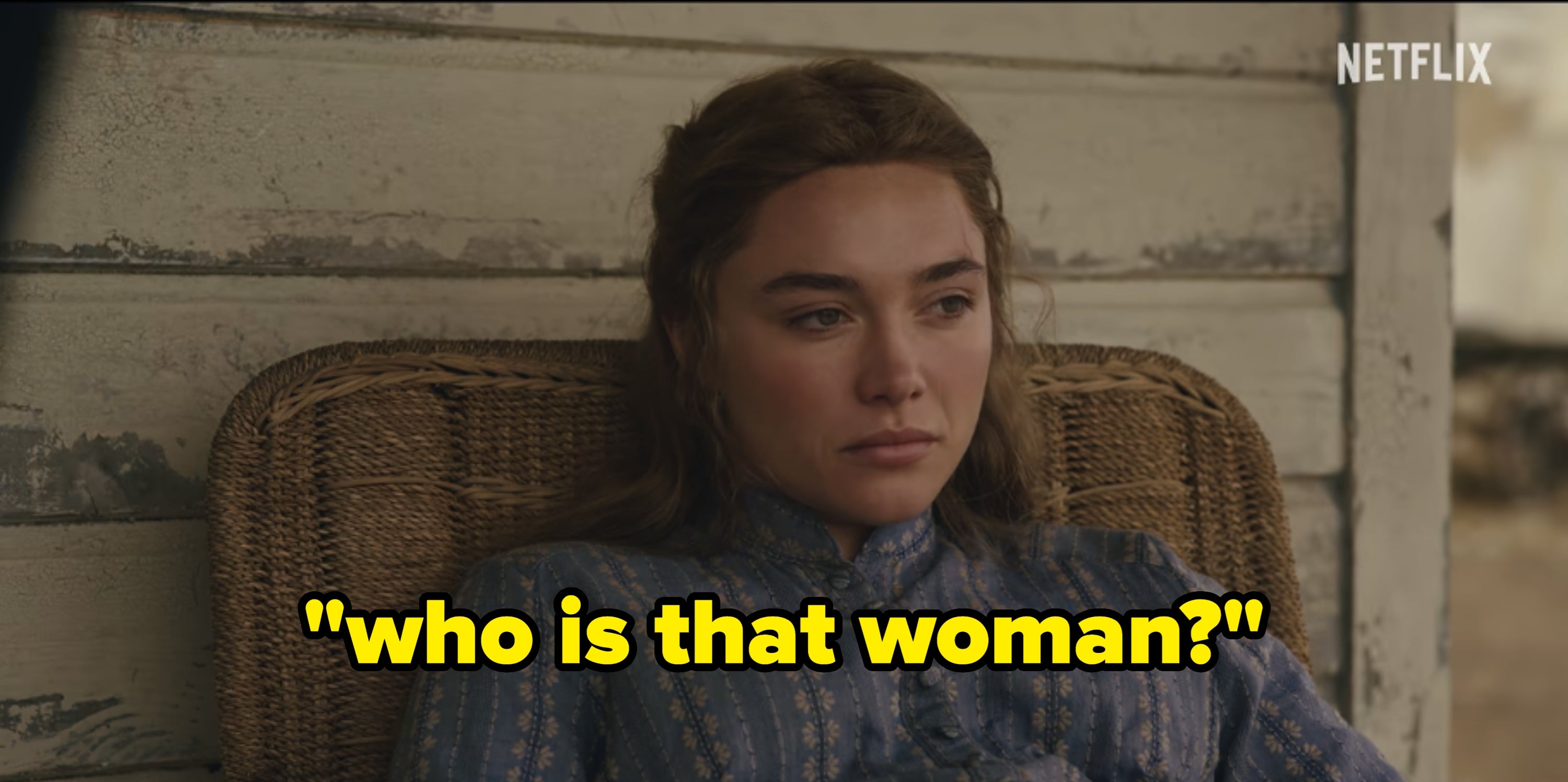 Person seated in a wicker chair wearing a vintage-style dress with a high collar, in a scene from a period film or TV show