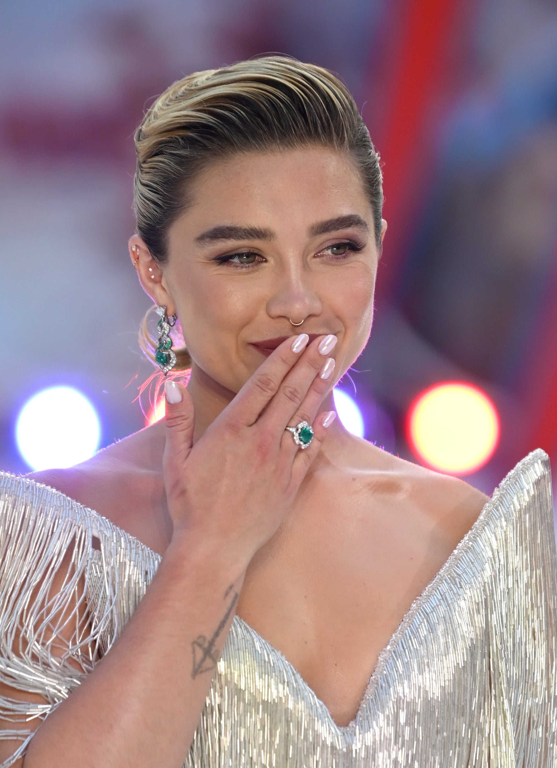 Person in a shimmering, v-neck gown with fringe details, wearing statement earrings and a ring, smiling with a hand over their mouth on the red carpet