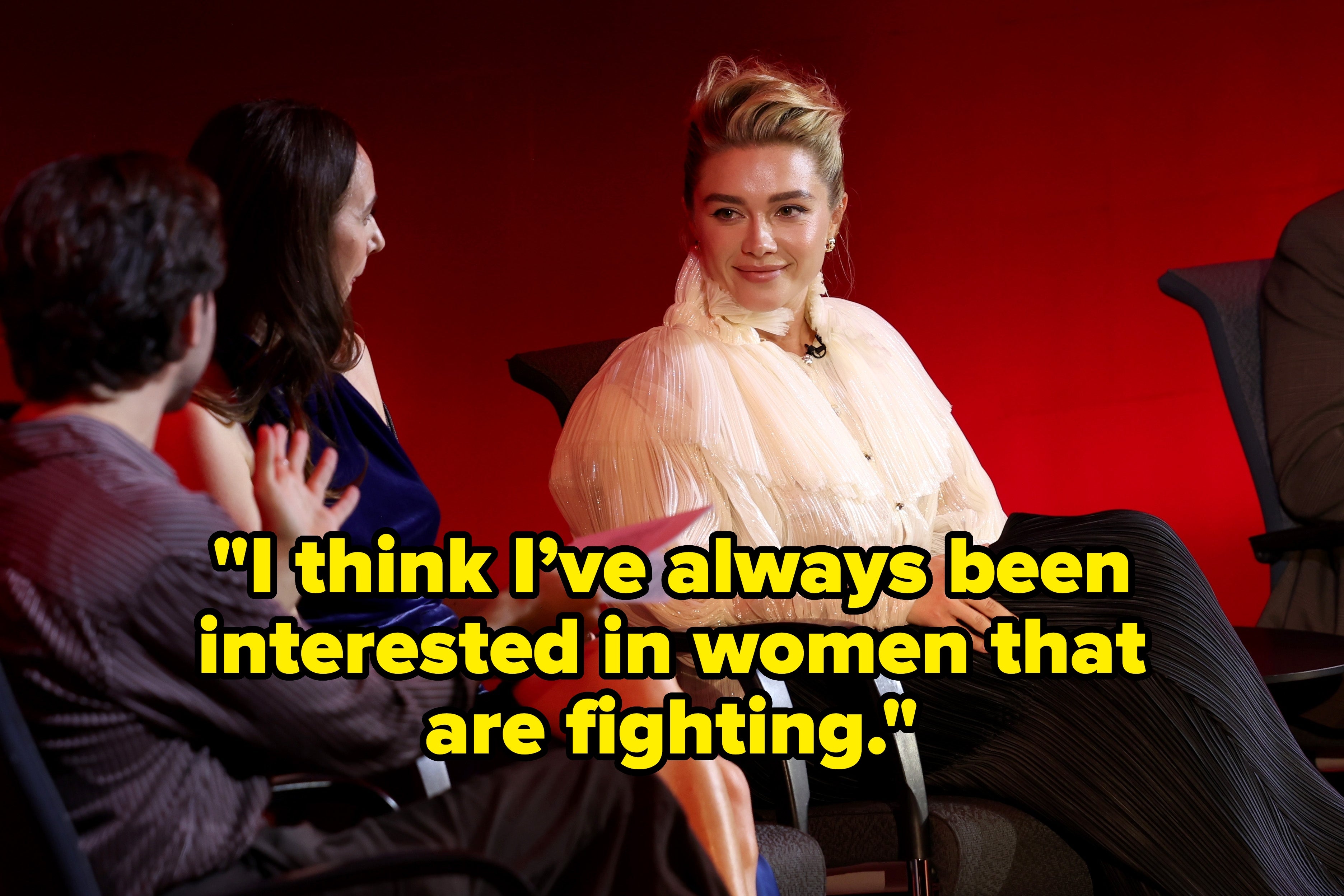 Person in elegant sheer blouse and pleated skirt sits on a panel with others, engaged in conversation against a red backdrop