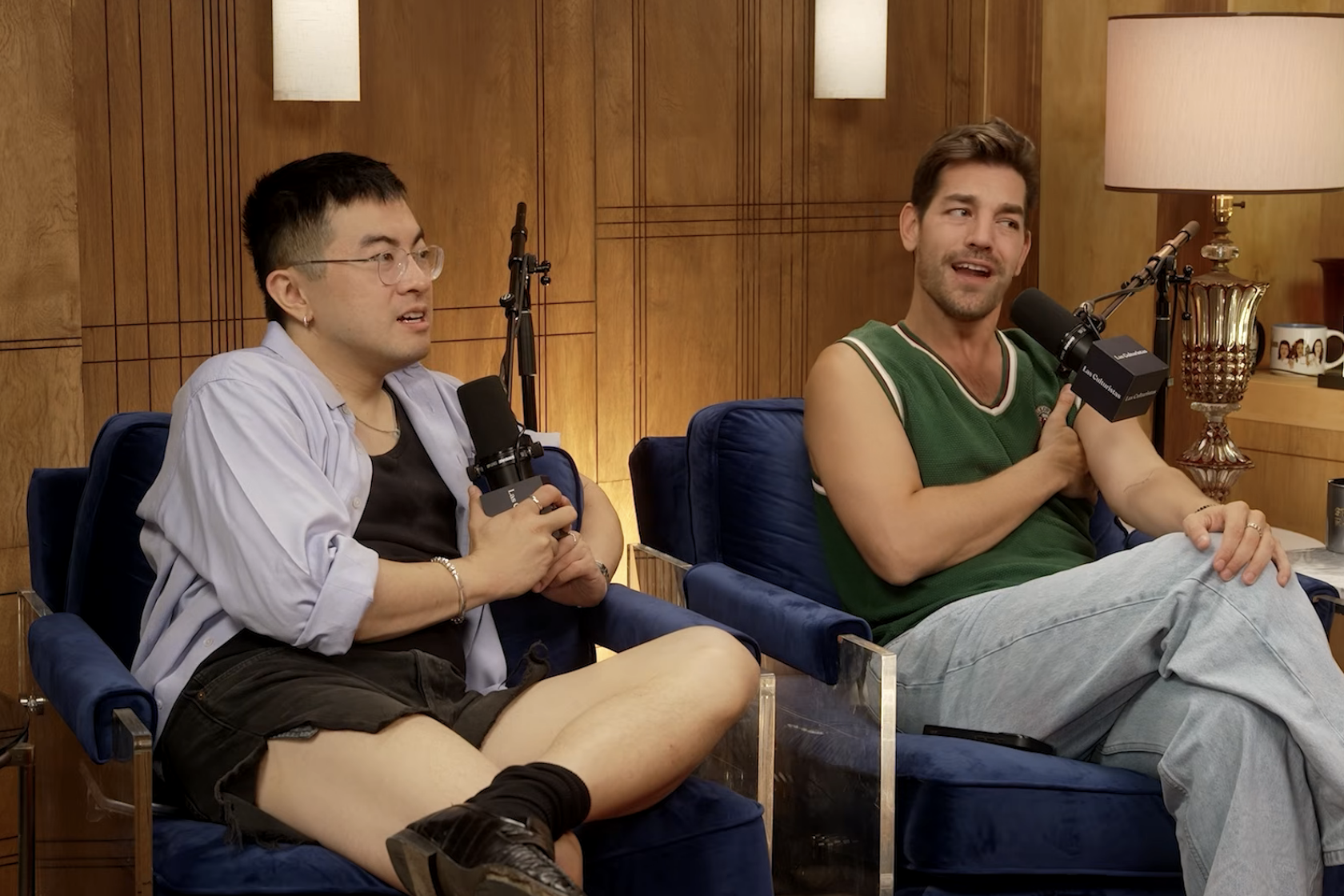 Two people sit and talk into microphones on a podcast set. One wears a casual outfit with a light jacket, the other a sleeveless top and jeans
