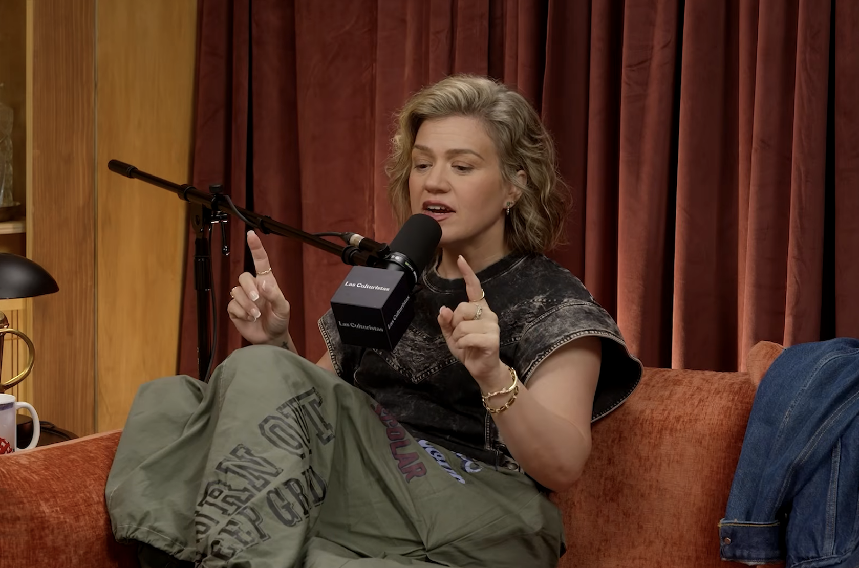 Person sitting on a sofa during a podcast interview, gesturing with both index fingers, and holding a microphone