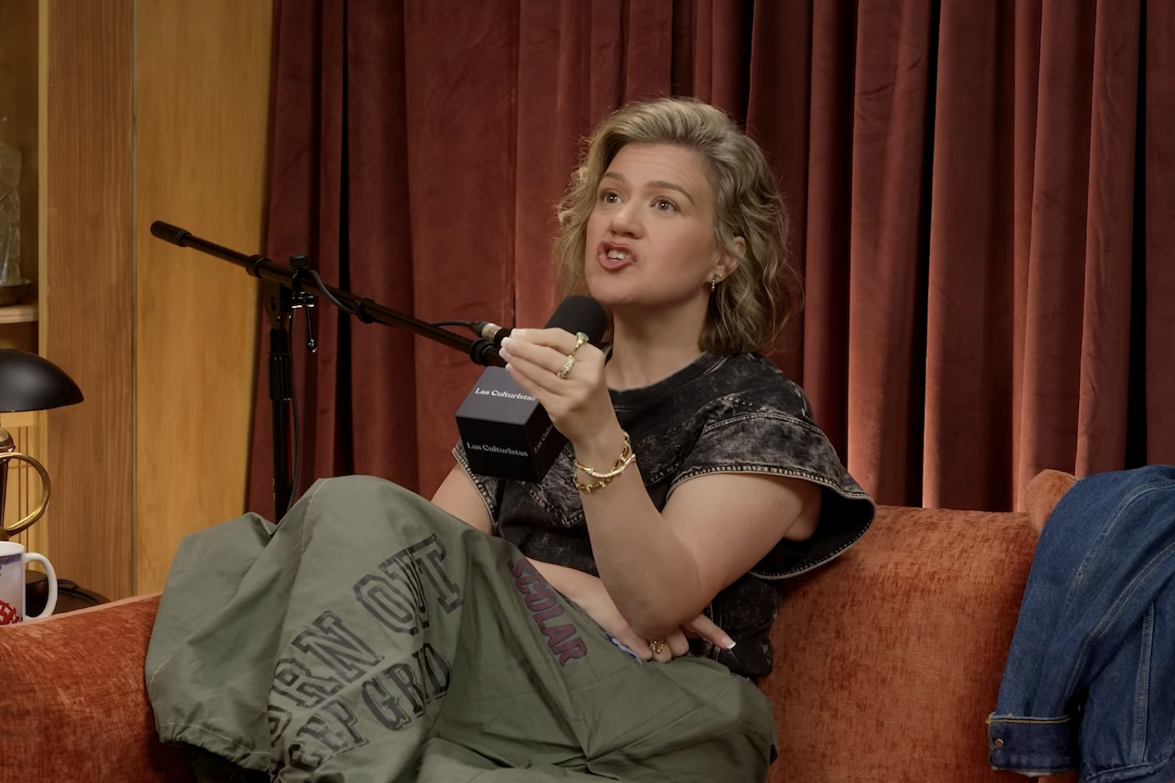 Person seated on a couch, speaking into a microphone. They are wearing a graphic T-shirt and baggy pants