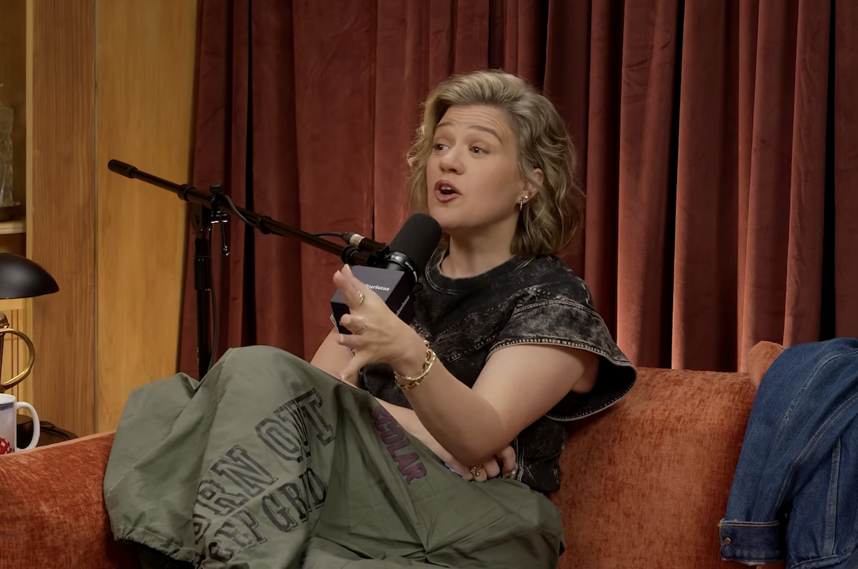 Person sitting on a couch, speaking into a microphone during a podcast or interview. Wearing casual, laid-back clothing