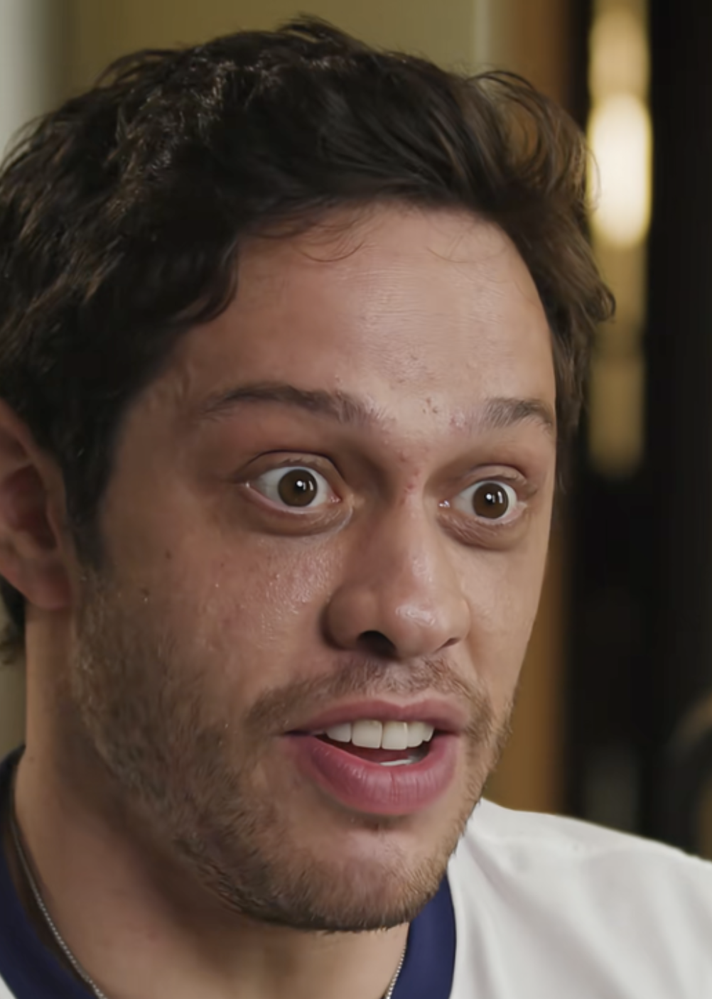 A person with wide eyes and an expressive face in a close-up shot, indoors, wearing a casual shirt
