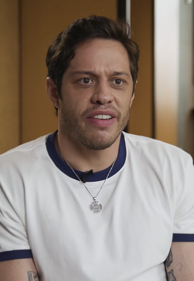 A person with short hair and facial hair wears a casual white T-shirt and necklace; they appear to be speaking or reacting