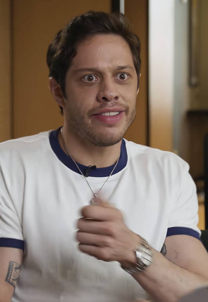 Person in a casual white T-shirt with a pendant necklace, gesturing during an animated conversation indoors