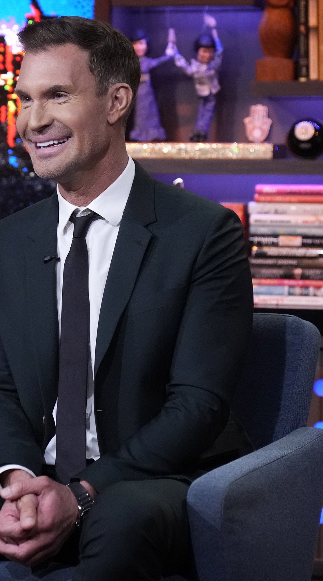 En person i dress sitter på et talkshow-sett og smiler. Bakgrunnen har bøker, en utstoppet leke og forskjellige dekorasjoner