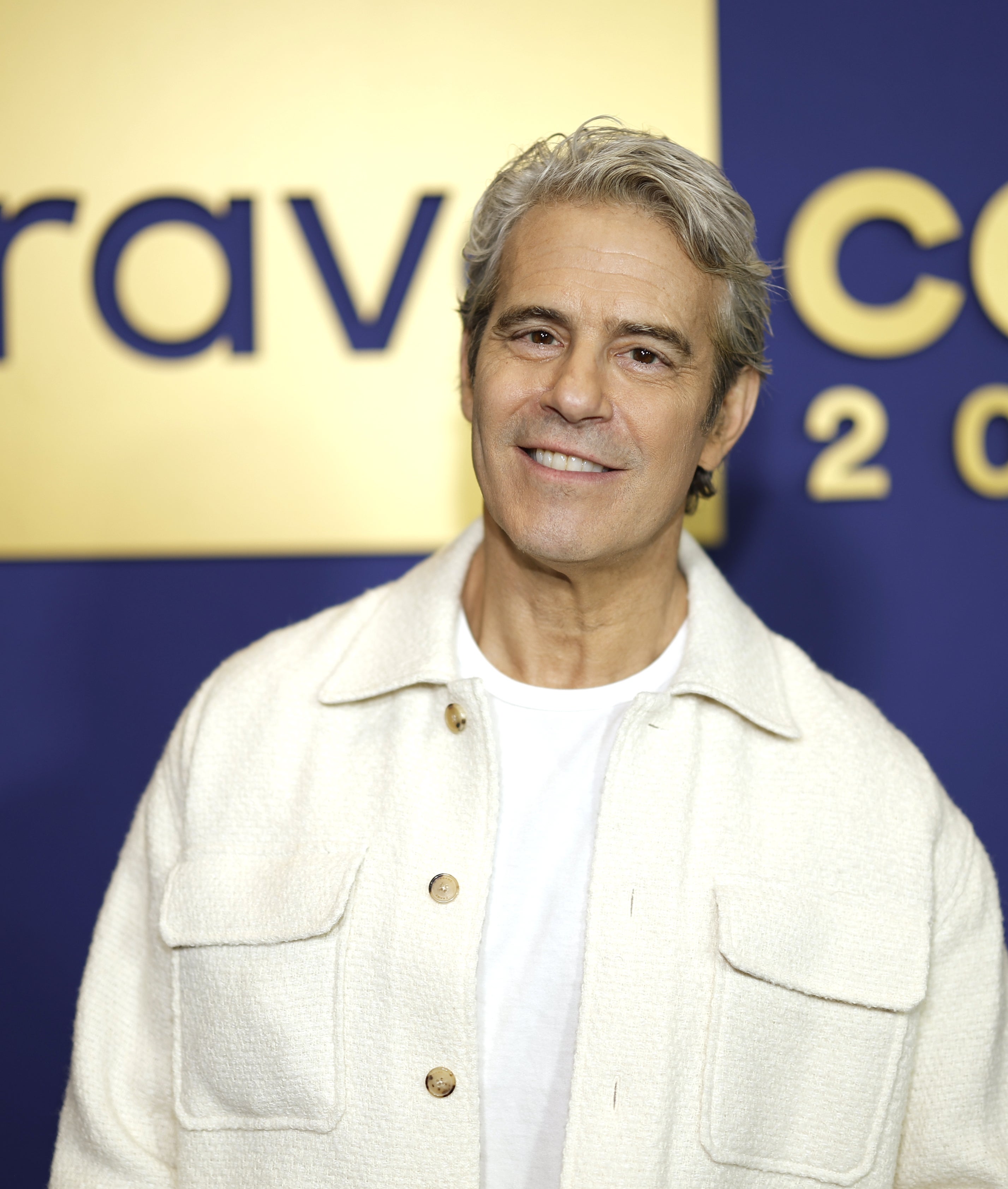 Andy Cohen i en hvit jakke smiler på bakgrunn av BravoCon 2025-arrangementet