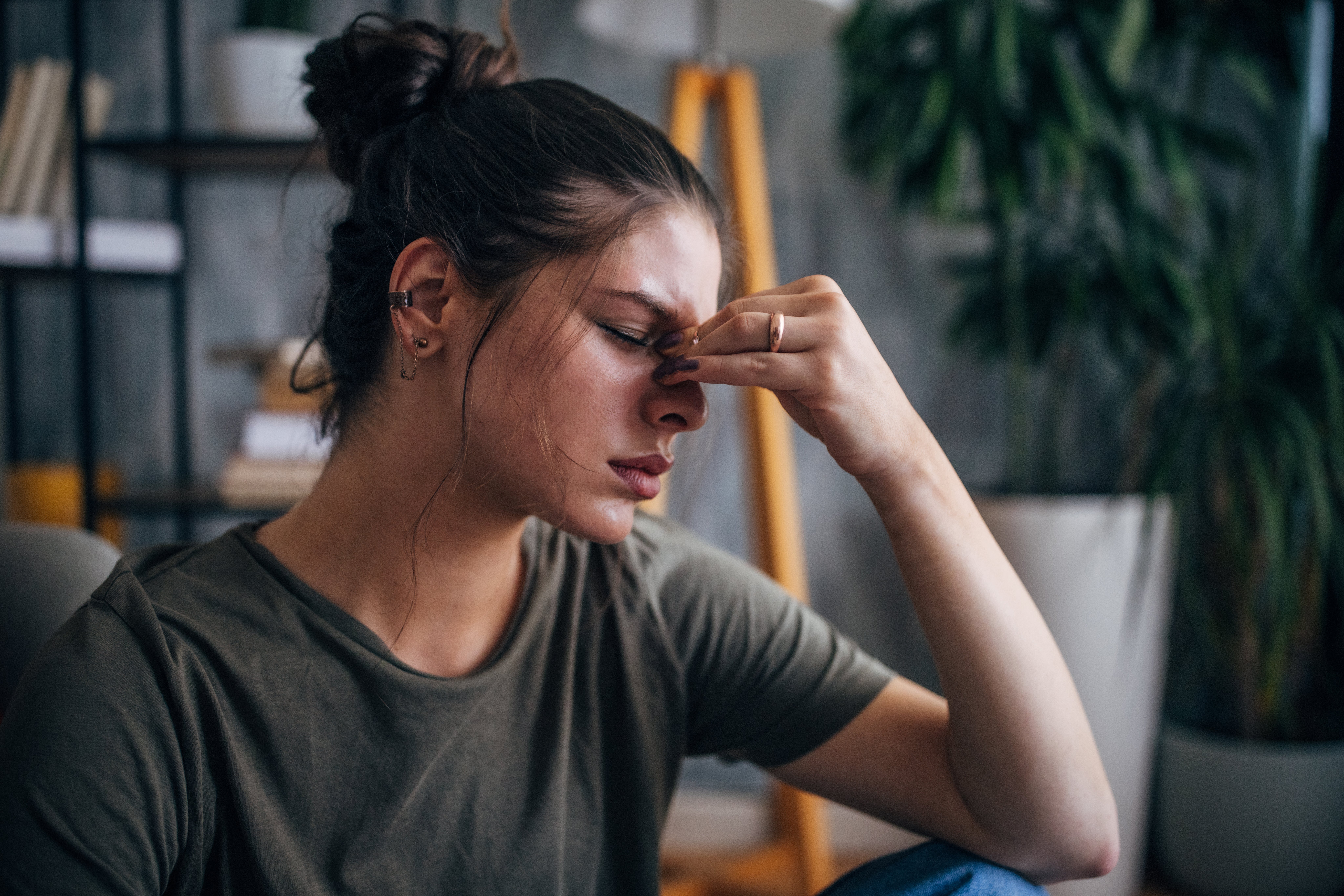 A person with closed eyes and a pained expression rubs their nose bridge, suggesting fatigue or stress, in a home setting